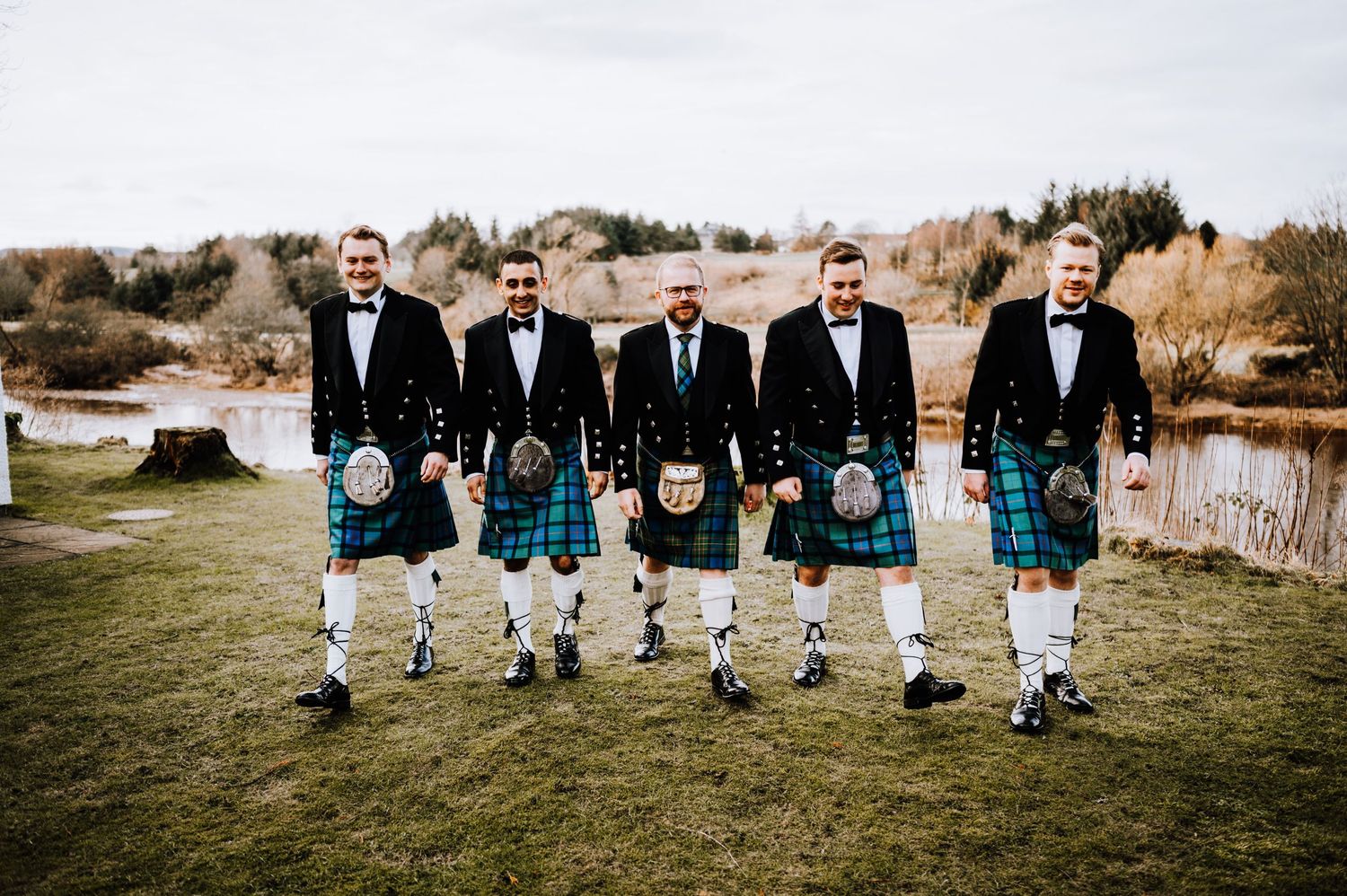 Five men in matching kilts and formal attire pose together on grass near a lake.