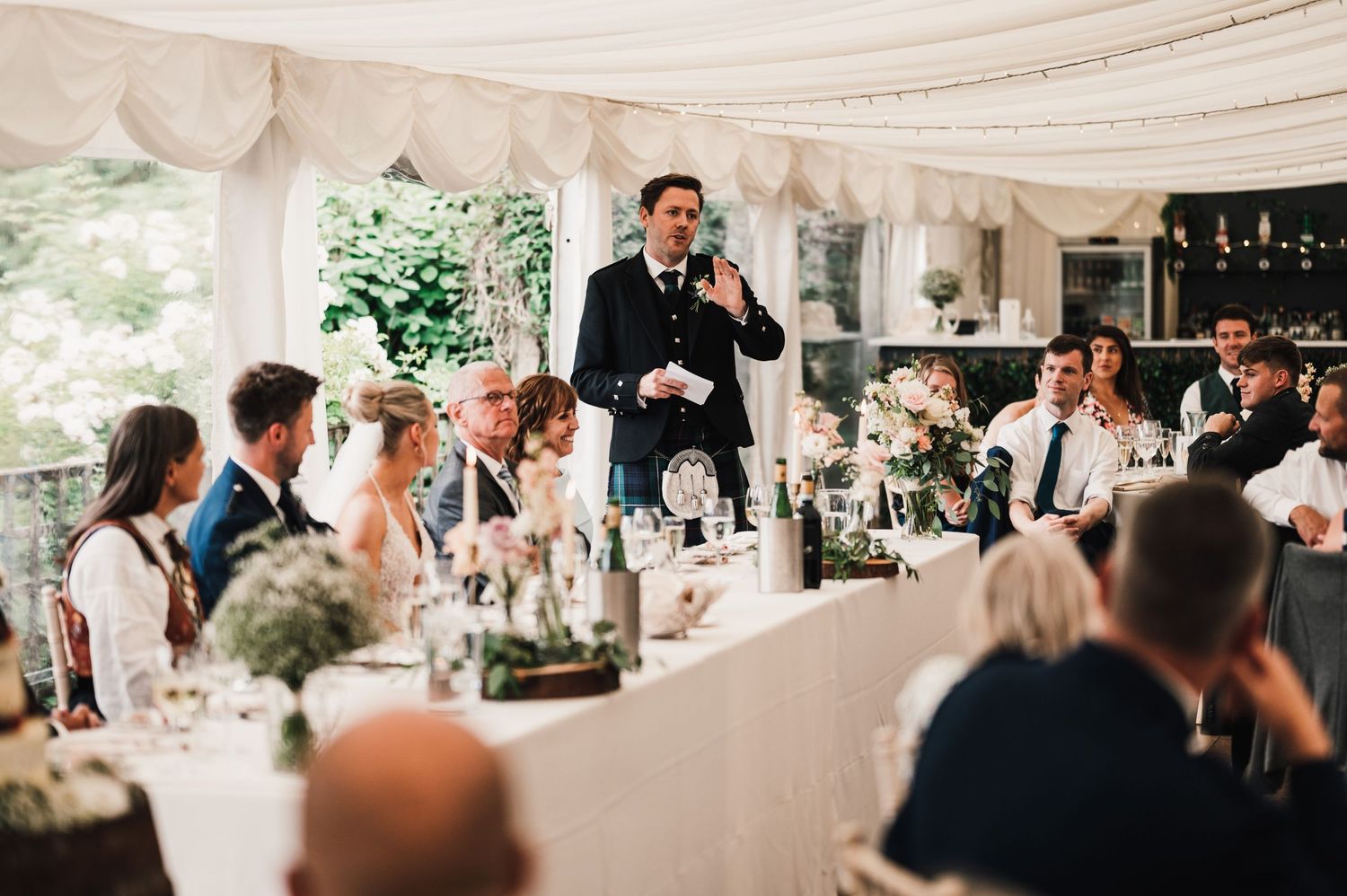 Wedding reception speech being delivered under elegant white marquee with guests listening attentively.