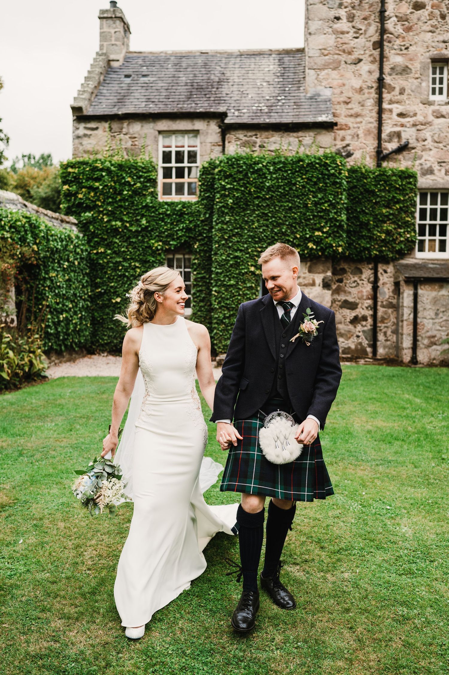 Joyful couple walking on lawn in front of ivy-covered Scottish stone house.