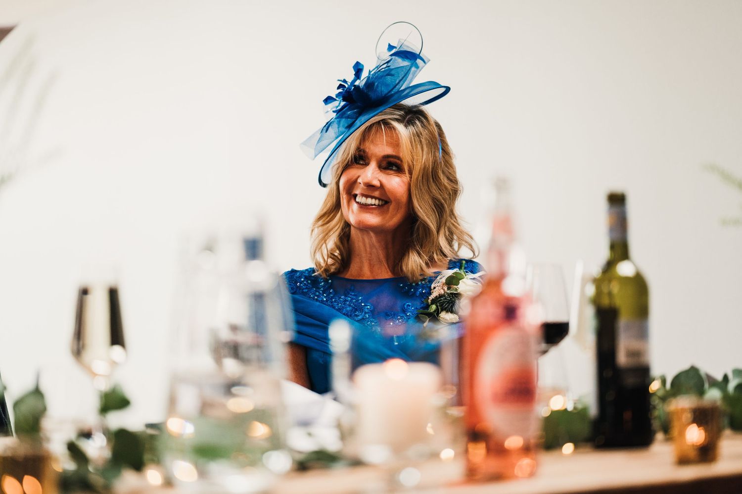 A guest in a blue fascinator enjoys a festive wedding reception with wine bottles and table settings.