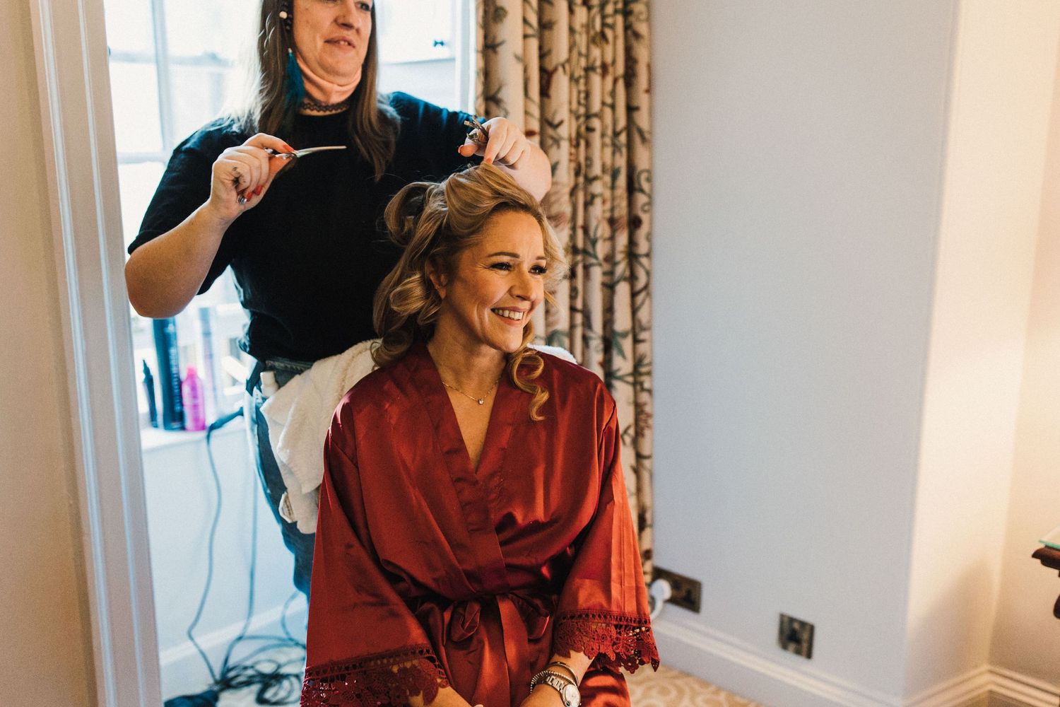 Wedding preparation scene with hairstyling session in a bright room with floral curtains.