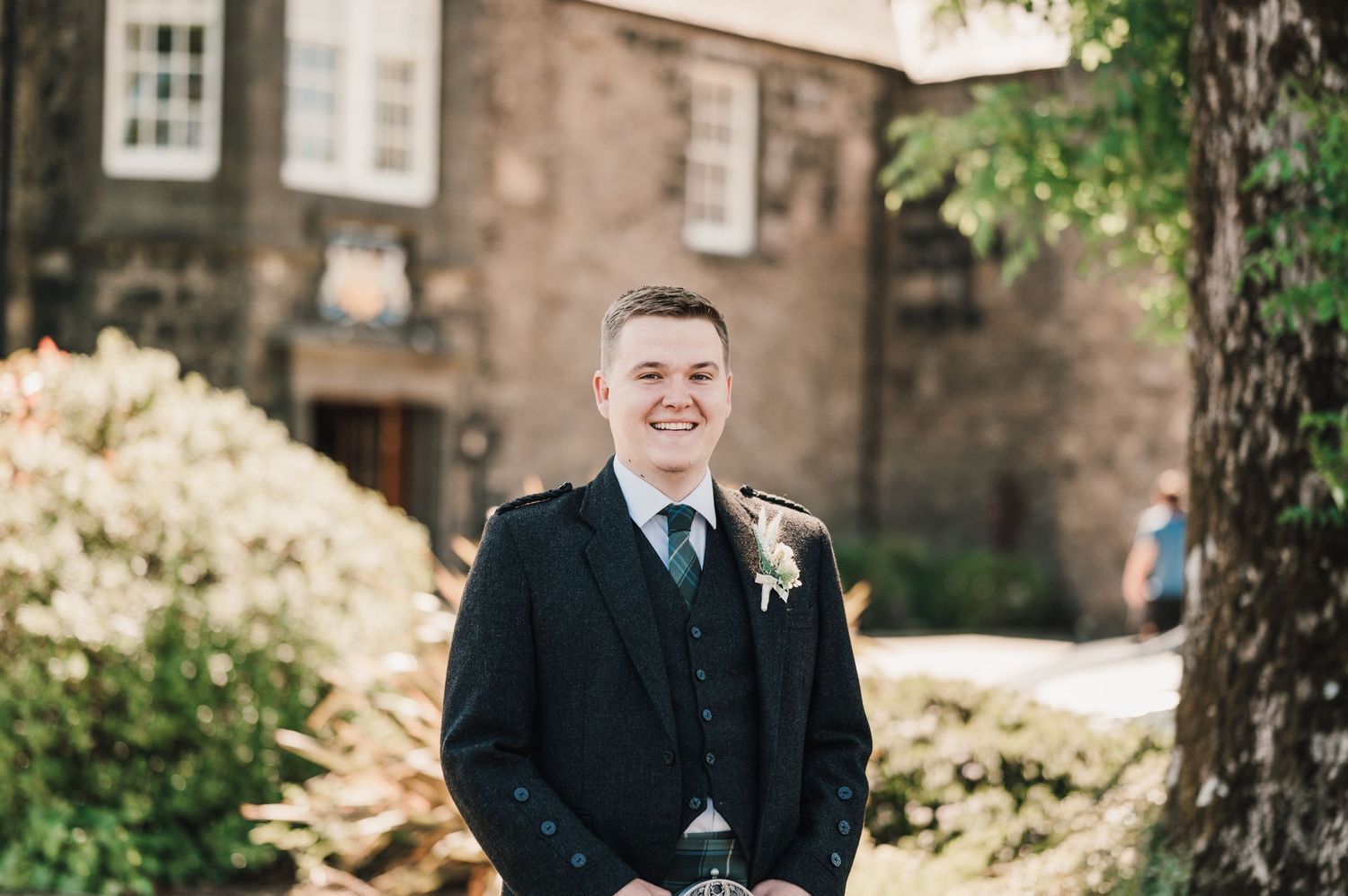 A person in traditional Scottish wedding attire smiles outside a historic stone building.
