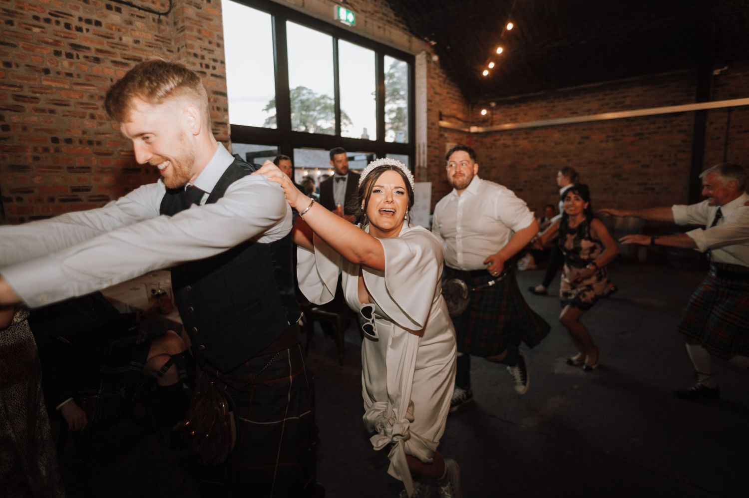 People dancing energetically at a wedding celebration in an industrial-style venue with brick walls.