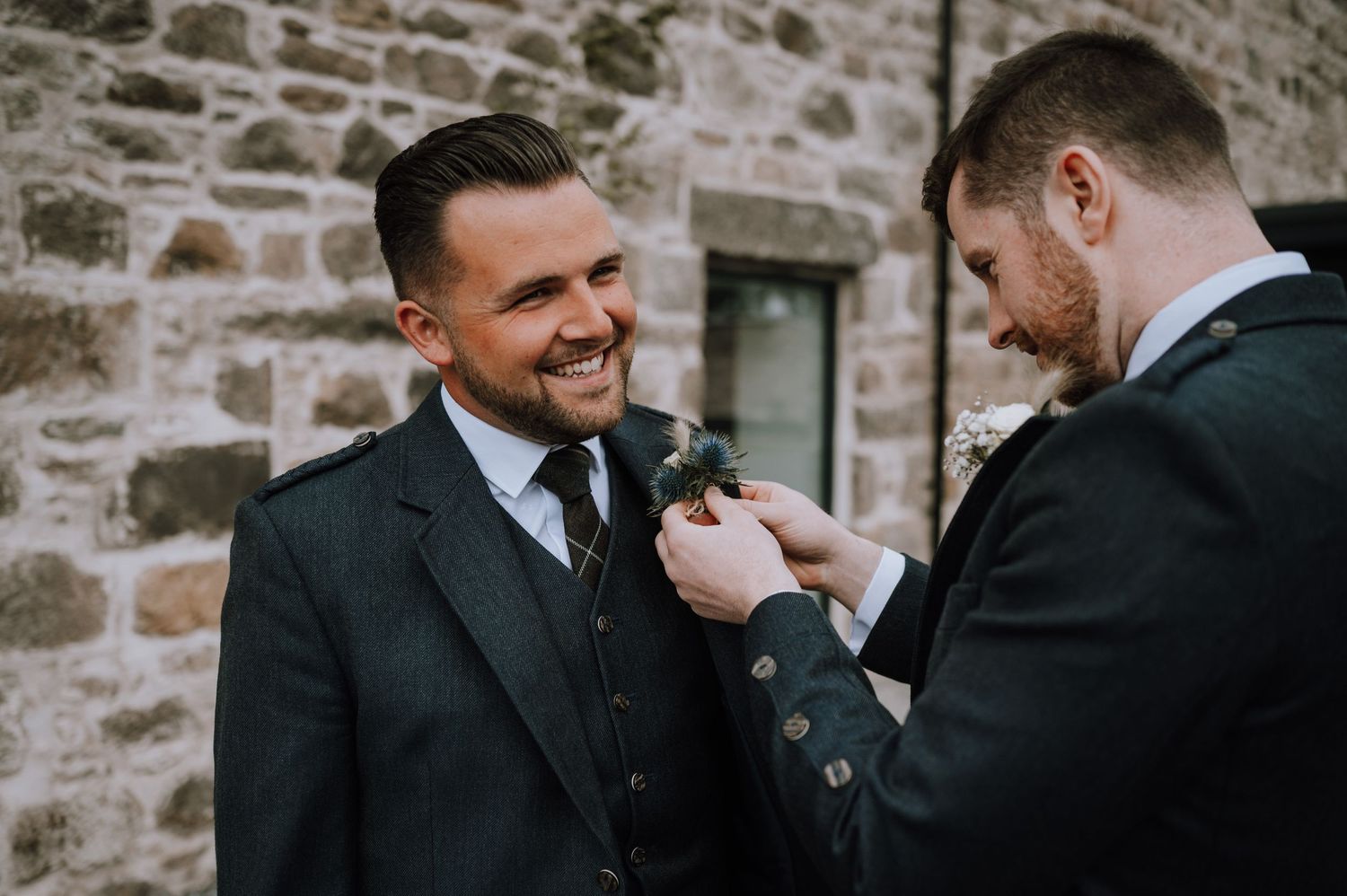 Two men in dark suits share a lighthearted moment while adjusting formal attire.