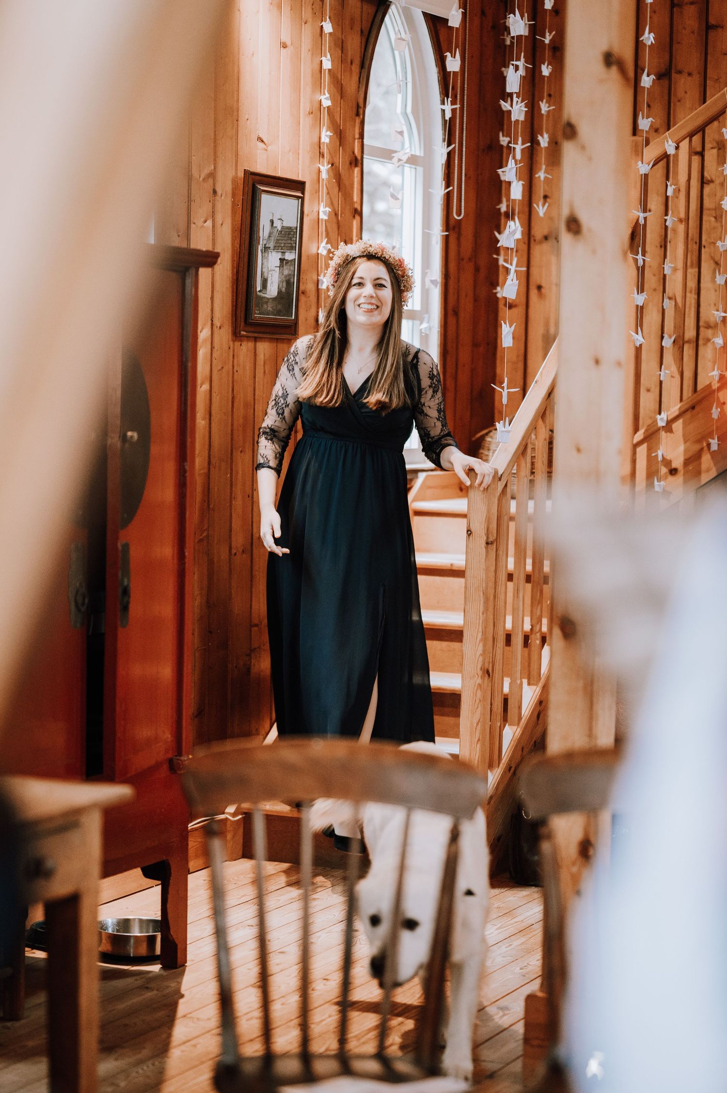 A person in a black dress stands on wooden stairs decorated with fairy lights in a rustic interior.