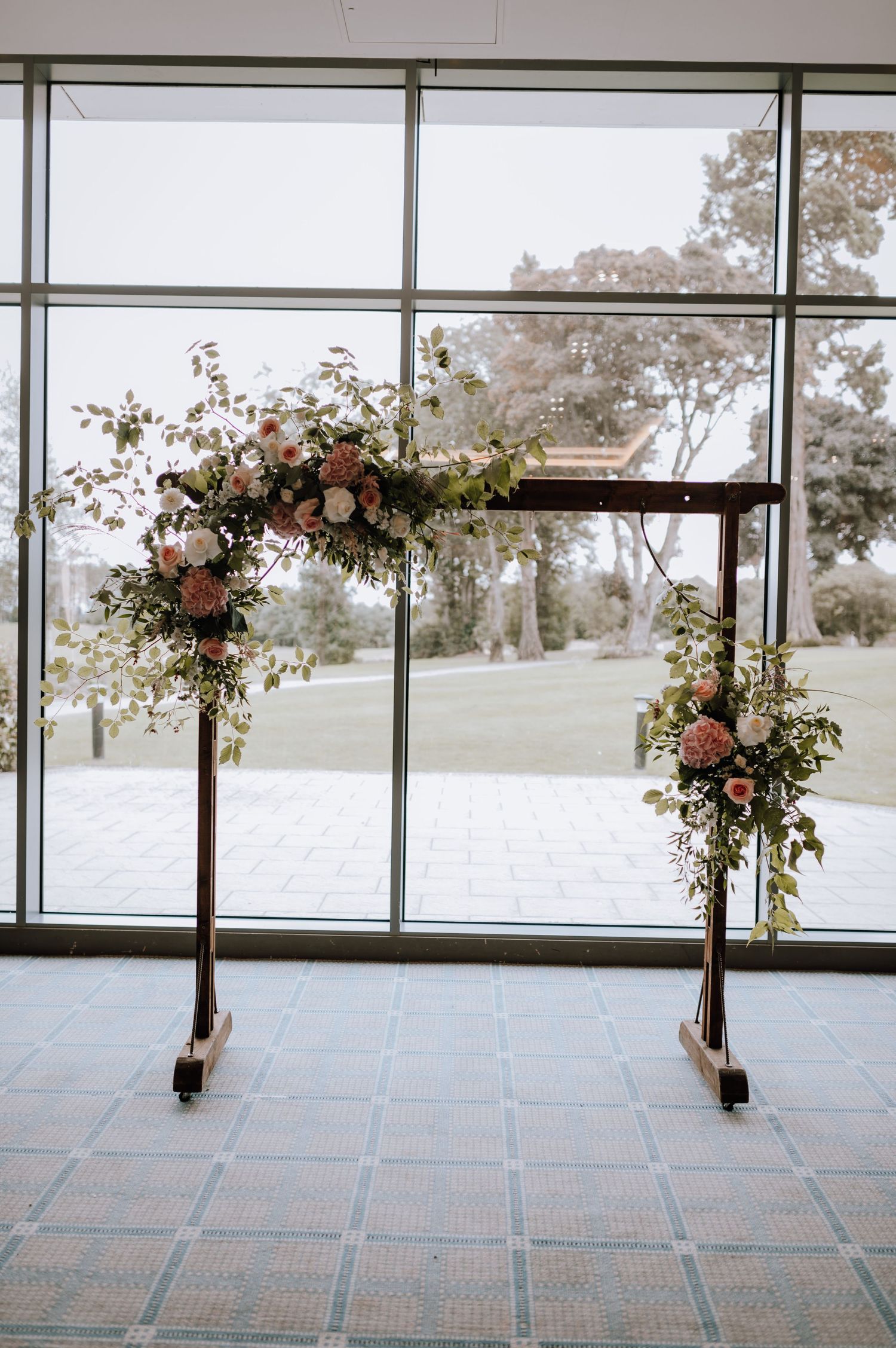 Wooden wedding arch decorated with pink roses and greenery stands against large windows overlooking an outdoor landscape.