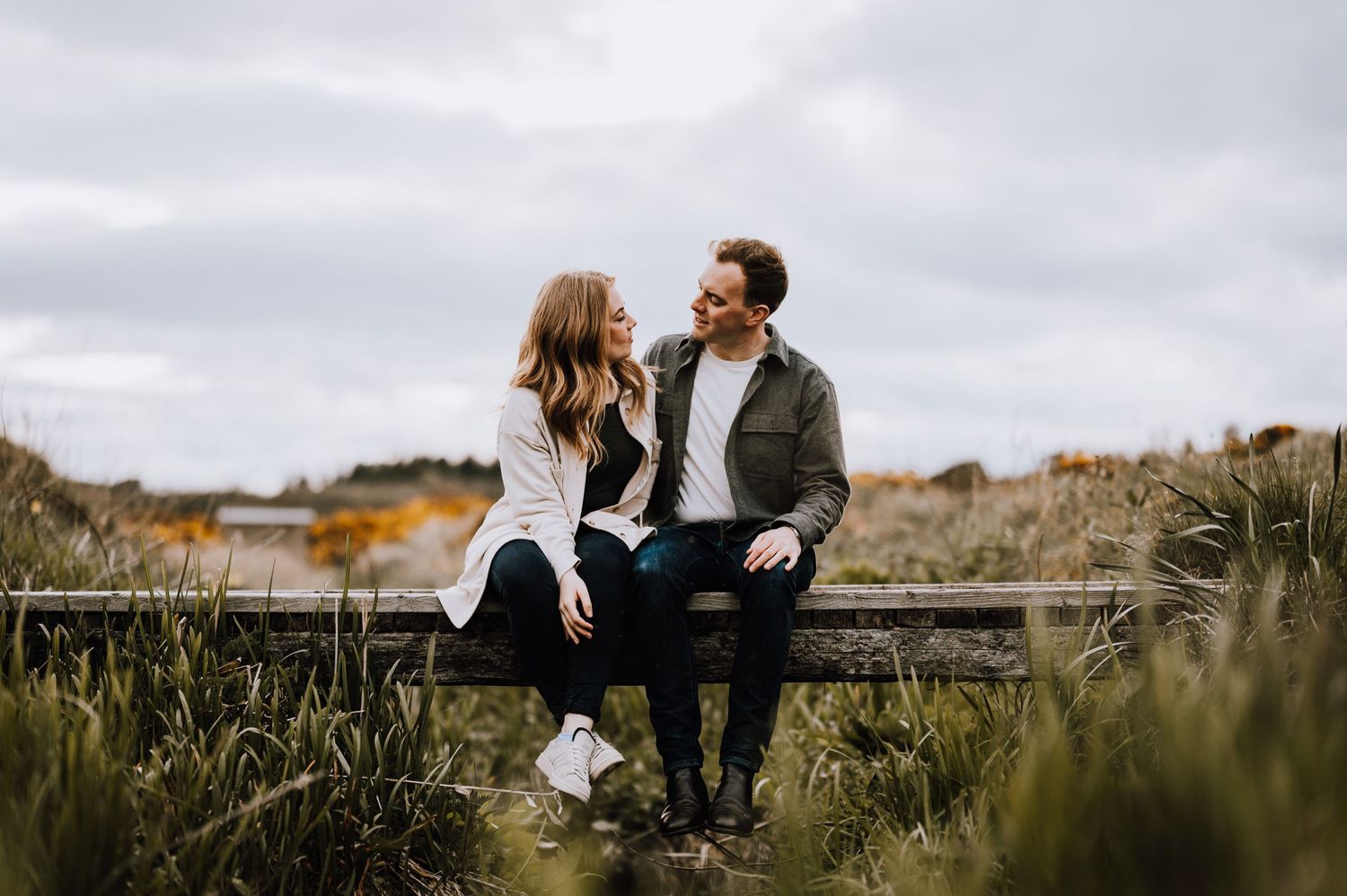 A couple sits casually on a wooden fence surrounded by wild coastal vegetation.