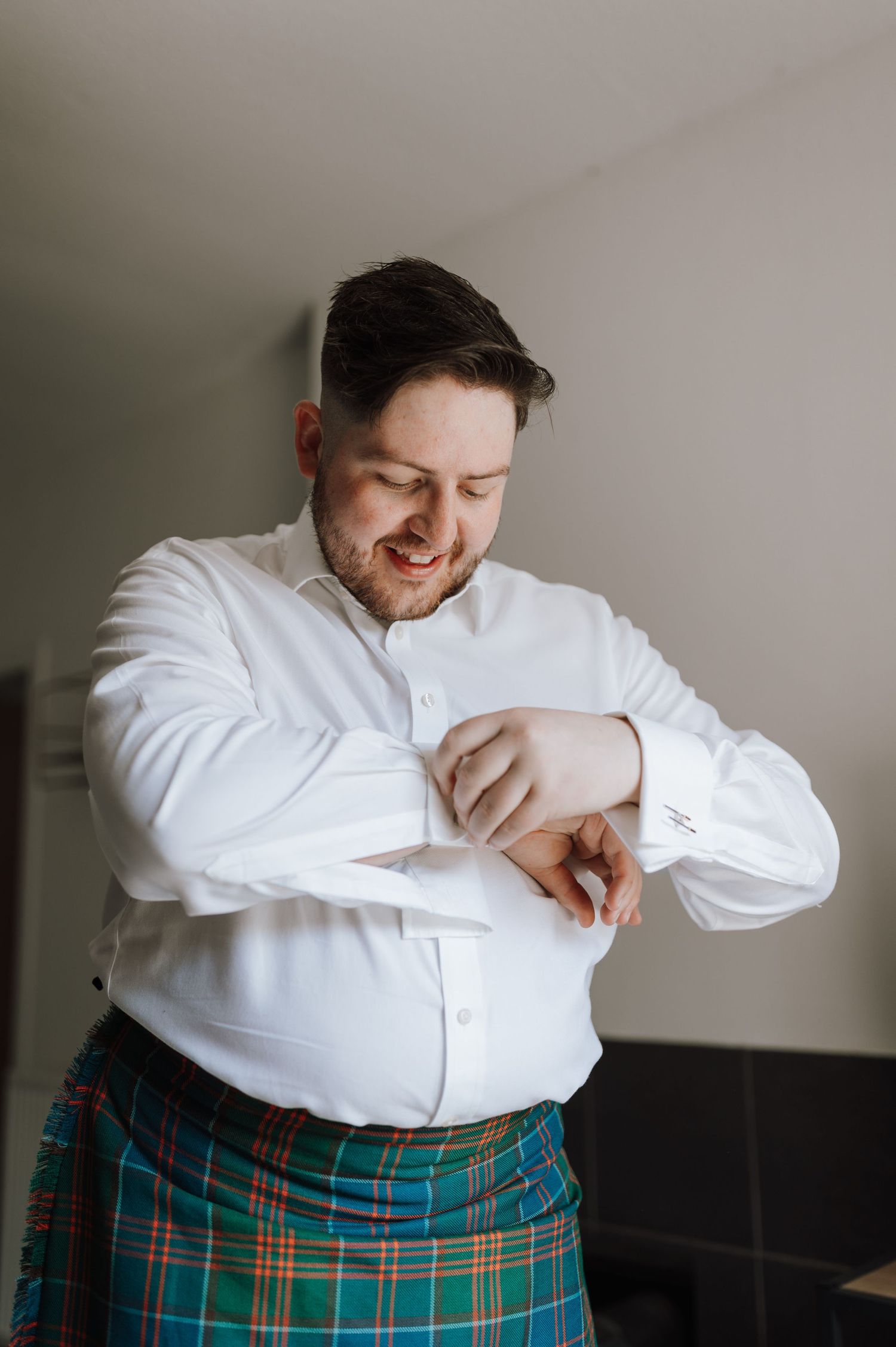 Person wearing traditional Scottish kilt and white dress shirt smiling while getting ready.
