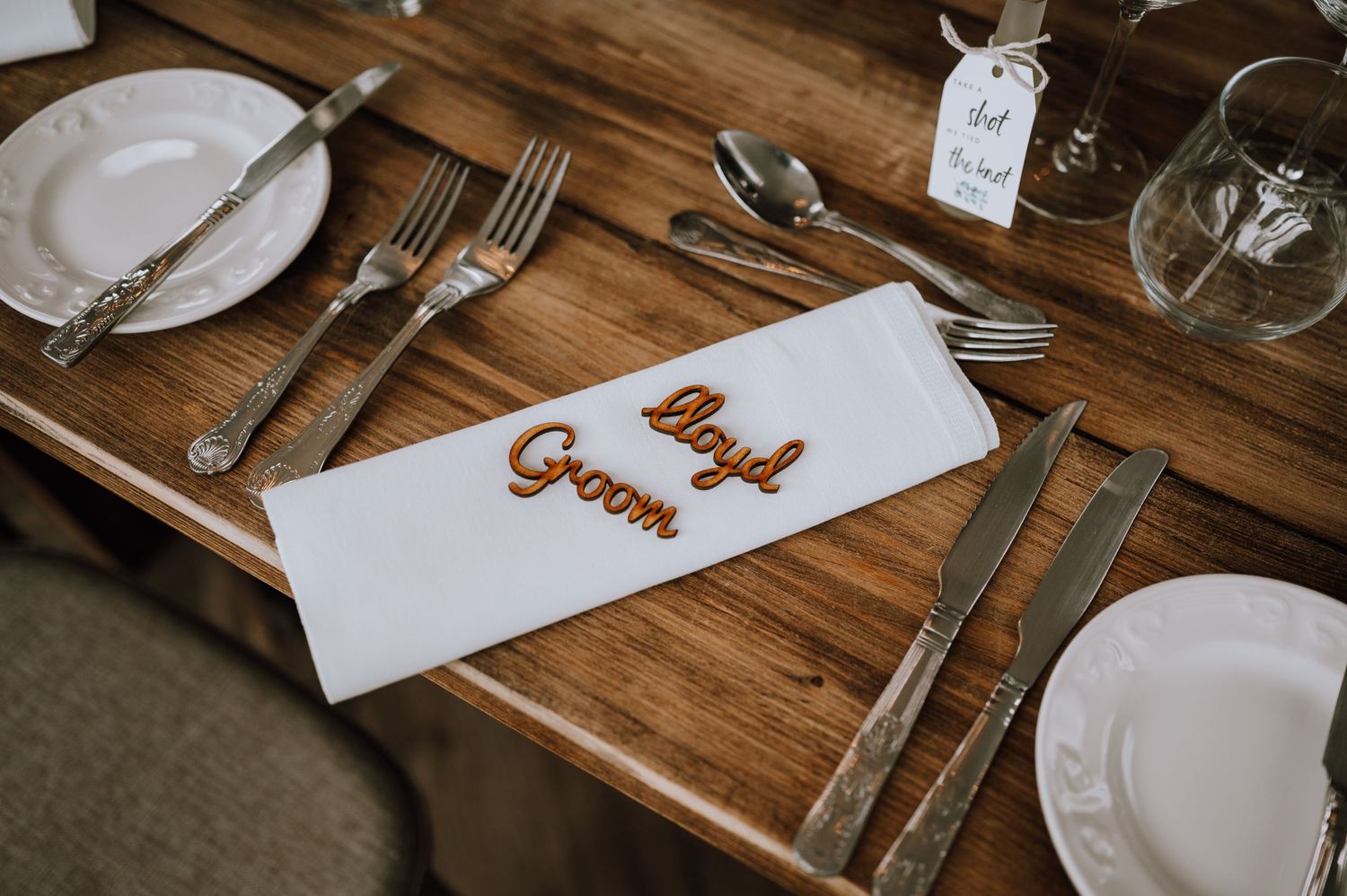 Place setting at a wedding reception with a white napkin marked 'Groom' on a rustic wooden table.