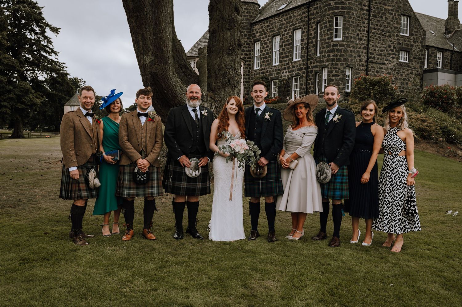 Wedding party members in kilts and formal wear pose for photo on castle lawn.