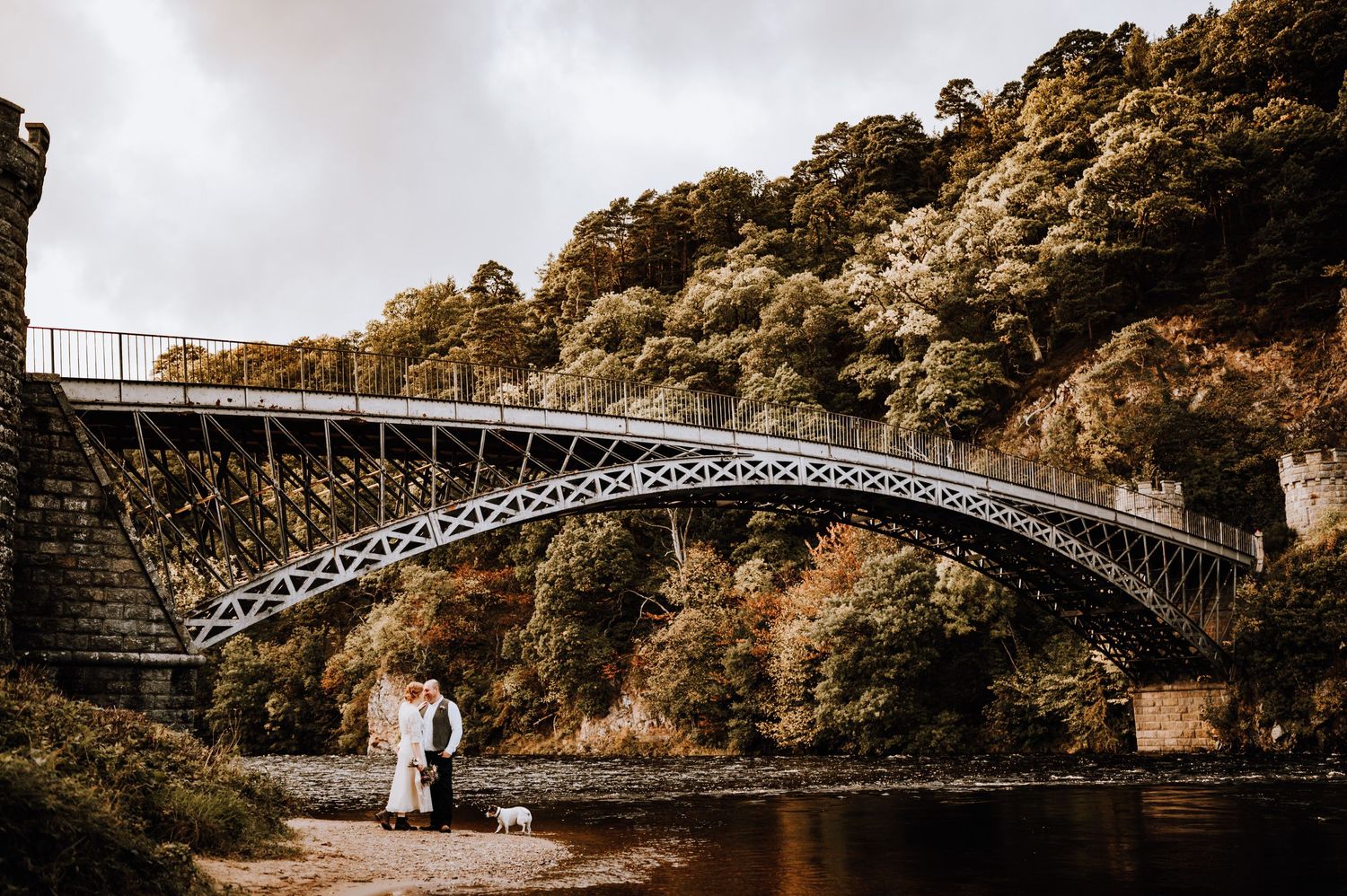 An ornate iron bridge spans across a river with lush autumn foliage on the surrounding cliffs.
