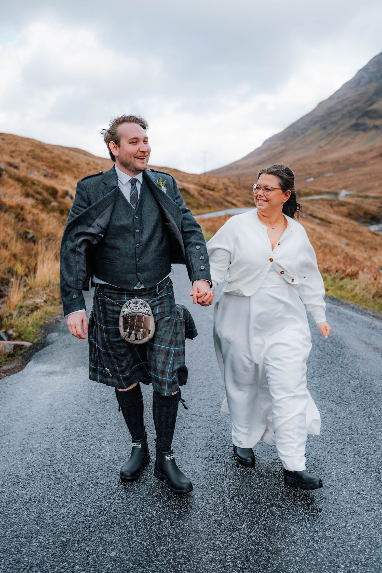 A couple walks together on a winding mountain road wearing traditional Scottish and white attire.