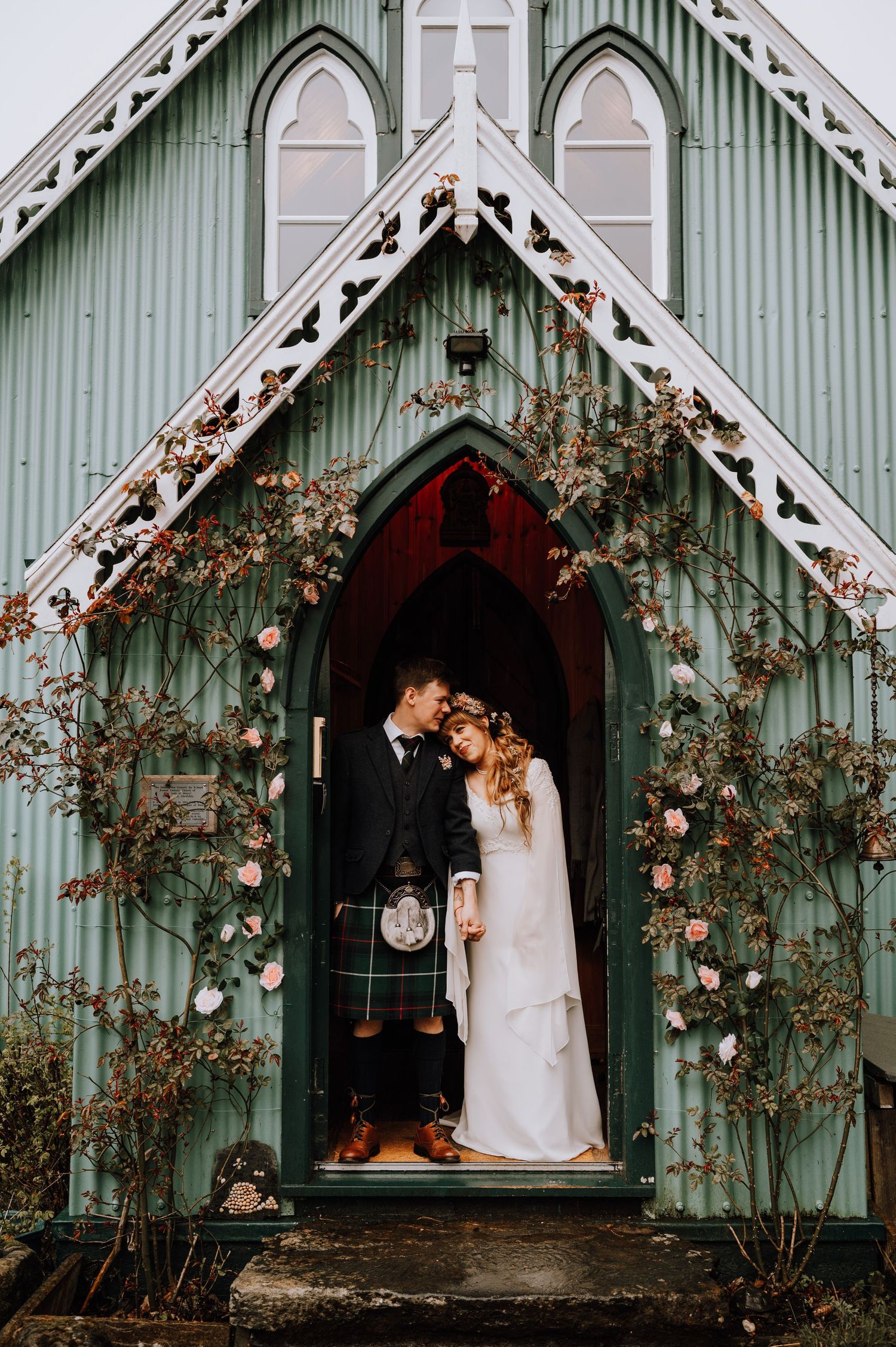 A Scottish wedding couple stands in the doorway of a mint green chapel adorned with climbing roses.