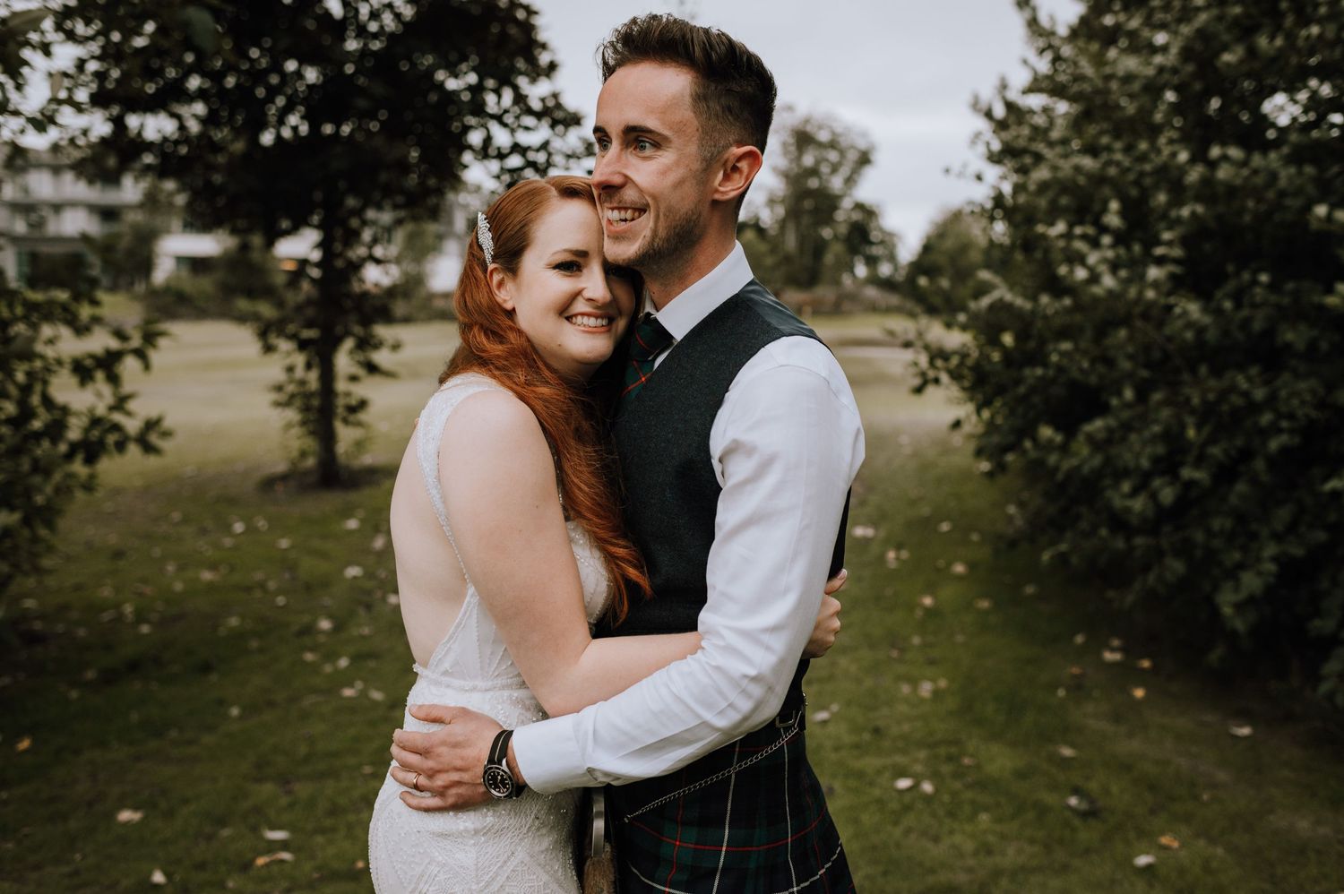 A couple in wedding attire embrace outdoors with the groom wearing traditional Scottish dress.