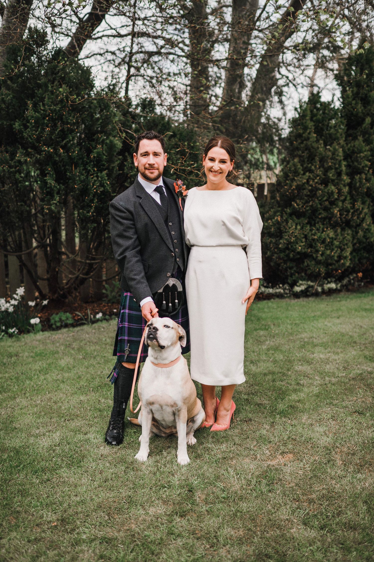 A couple poses with their dog in a garden, with the groom wearing traditional Scottish kilt attire.