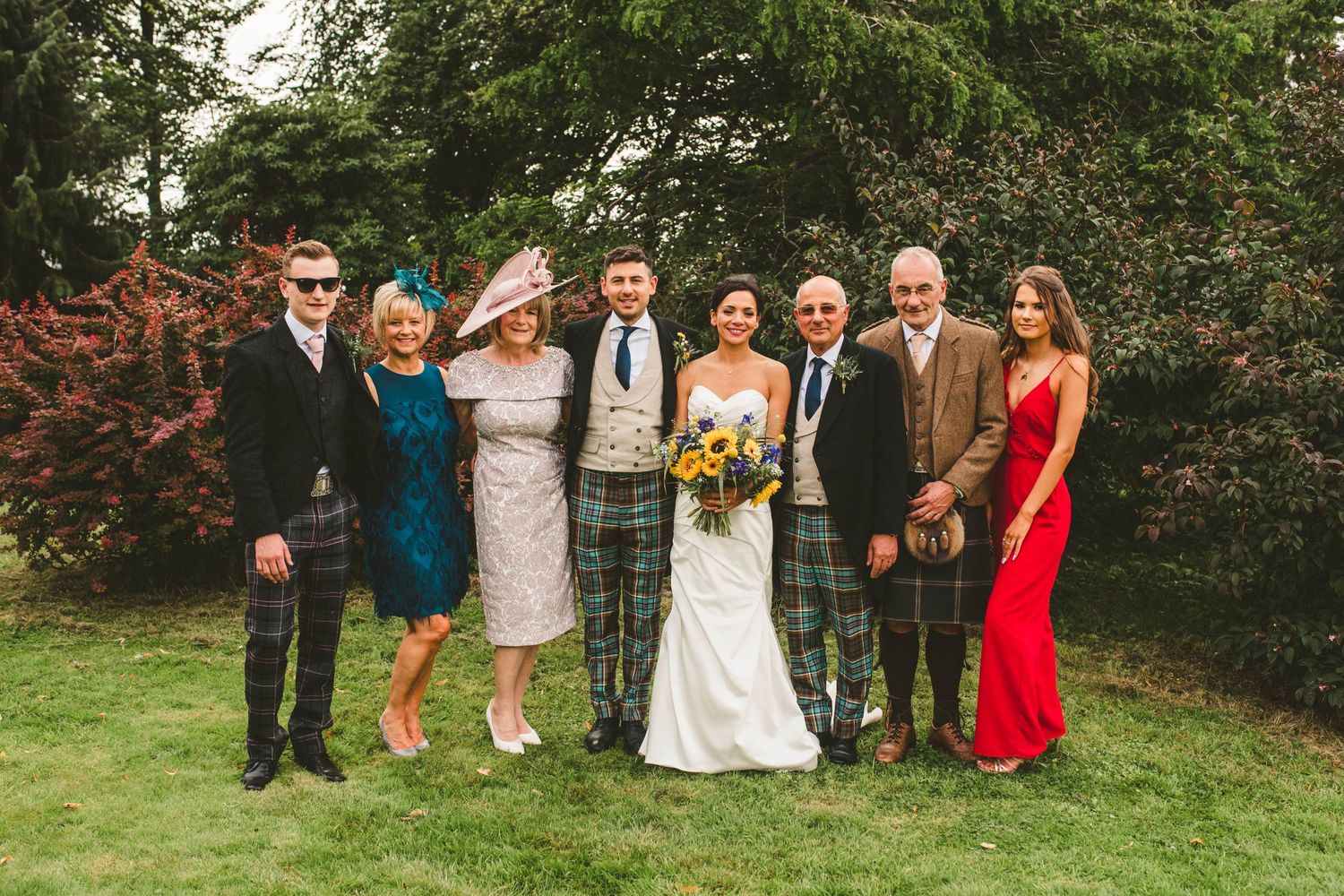 Wedding party in mix of kilts and formal attire pose together on garden lawn.