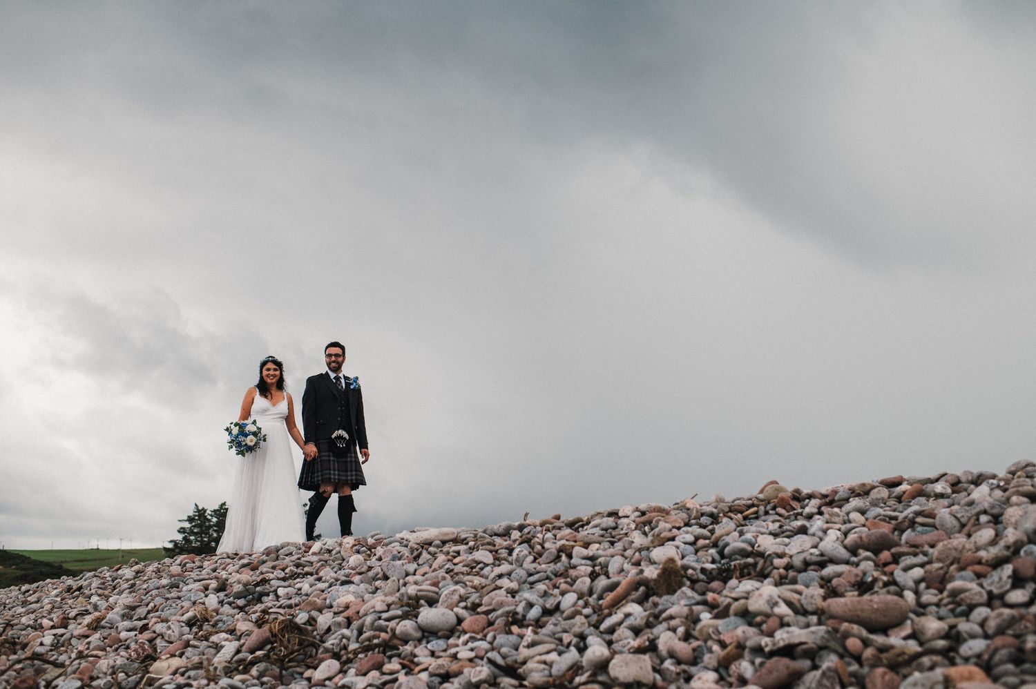 A wedding couple in Scottish attire stands on rocky terrain under stormy skies.