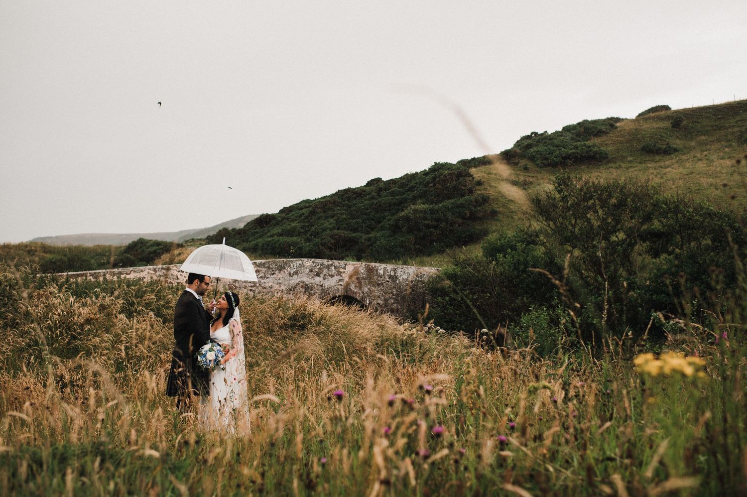 A couple in wedding attire stands with an umbrella in a misty field with rolling hills.