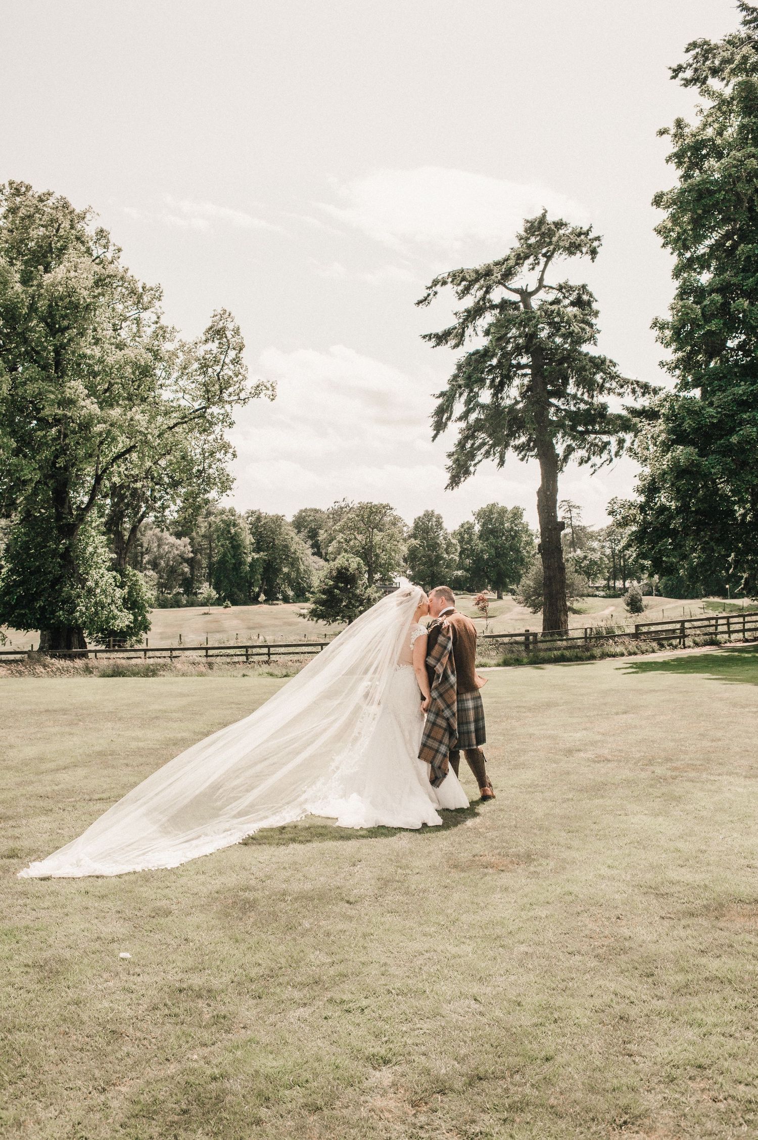 A romantic wedding couple embraces on manicured lawn with flowing veil and traditional Scottish attire.