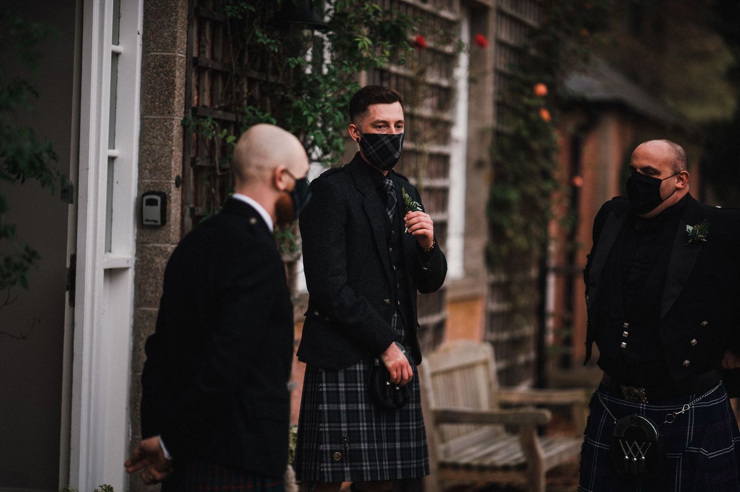 Group wearing kilts and dark jackets gathered outside brick building entrance.