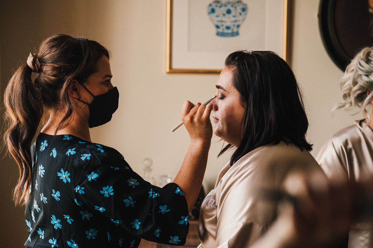 Two people in masks working on wedding makeup preparation in a softly lit room.
