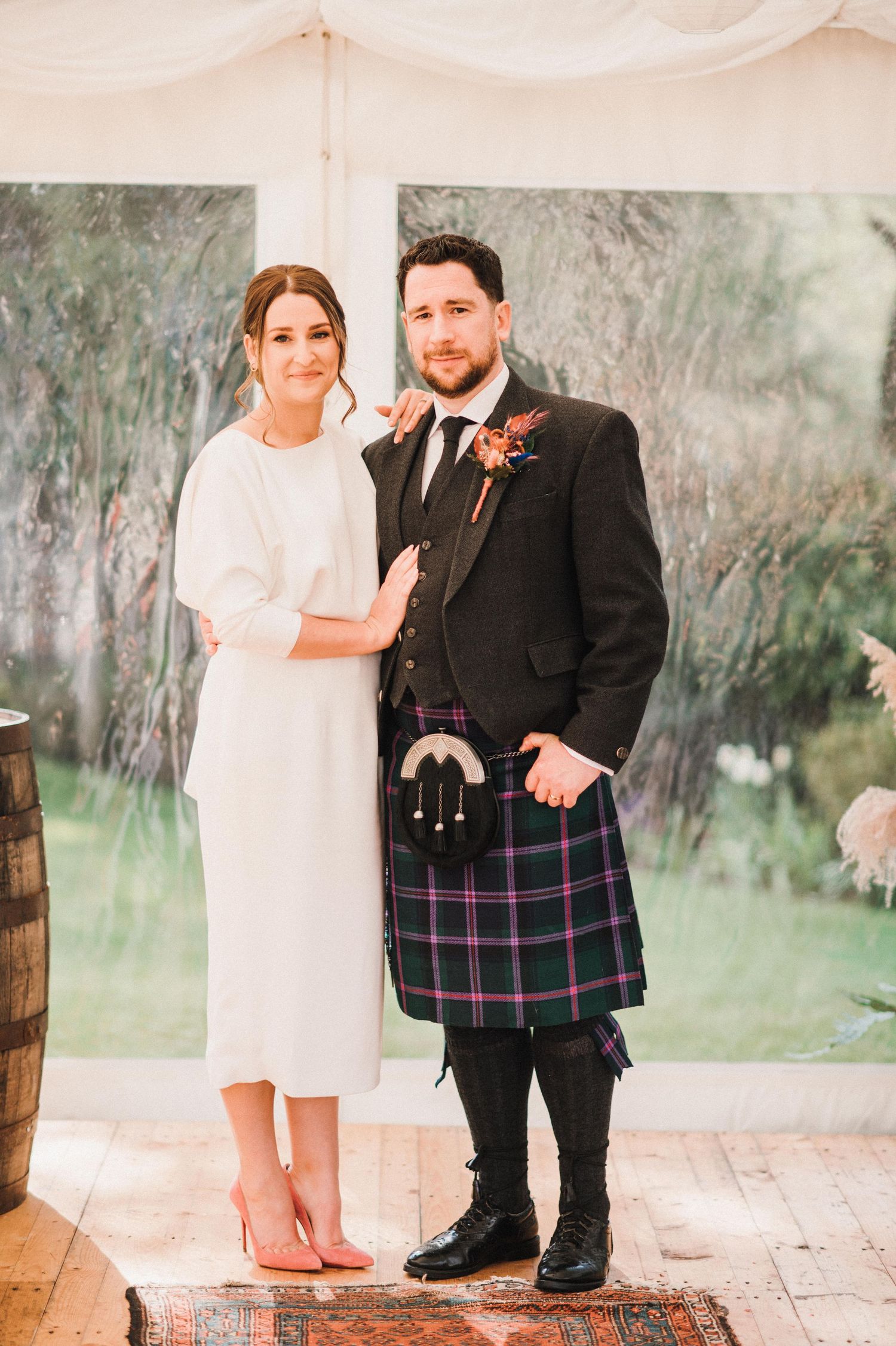 Newlyweds posing under white tent, groom wearing traditional Scottish kilt.