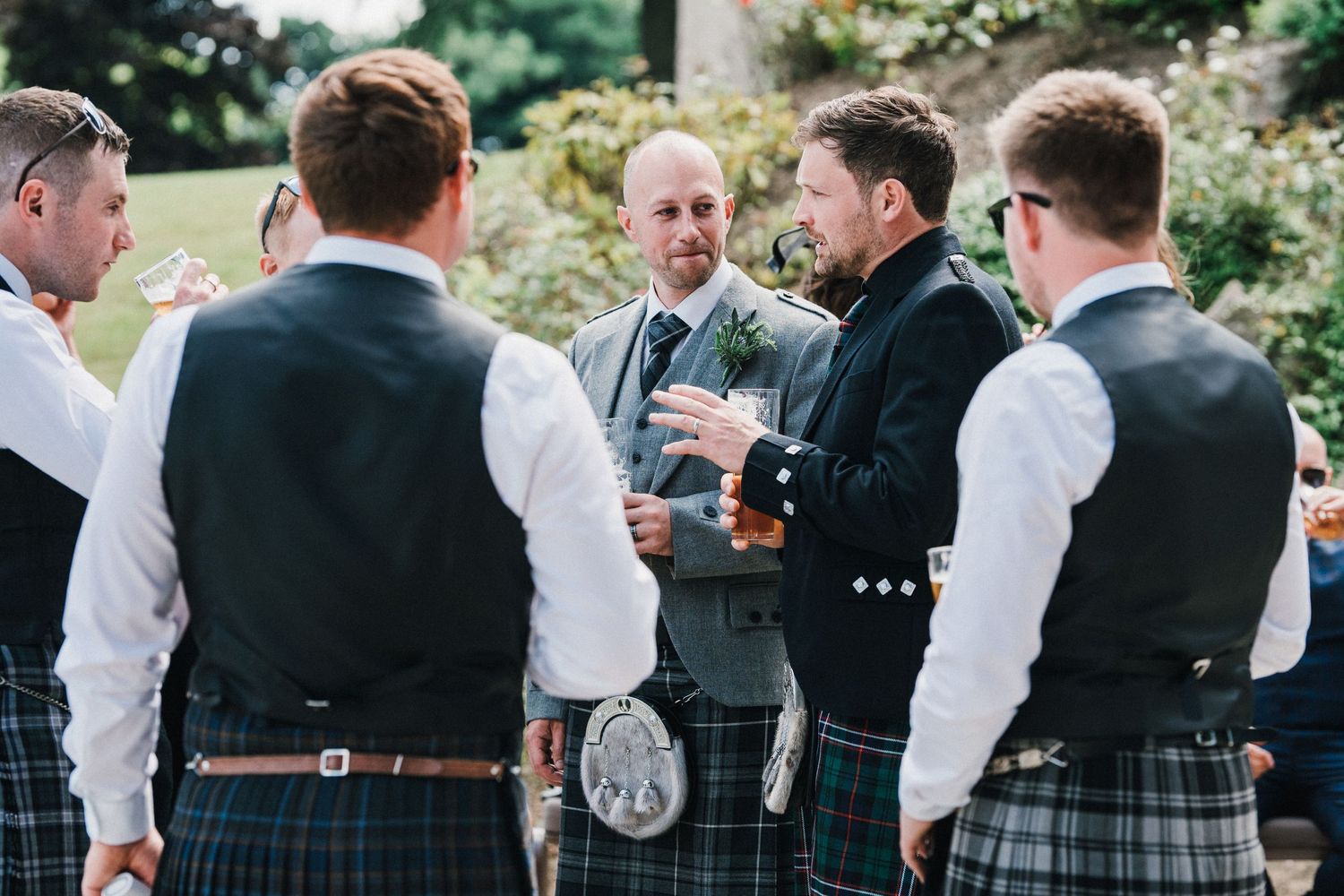 Group of men in kilts and formal attire gather for drinks at outdoor wedding reception.