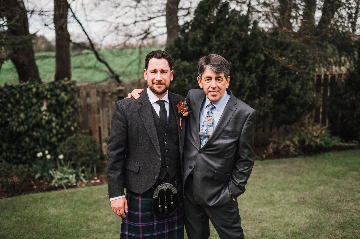 Wedding guests in formal attire pose together outdoors, with one wearing a traditional Scottish kilt.