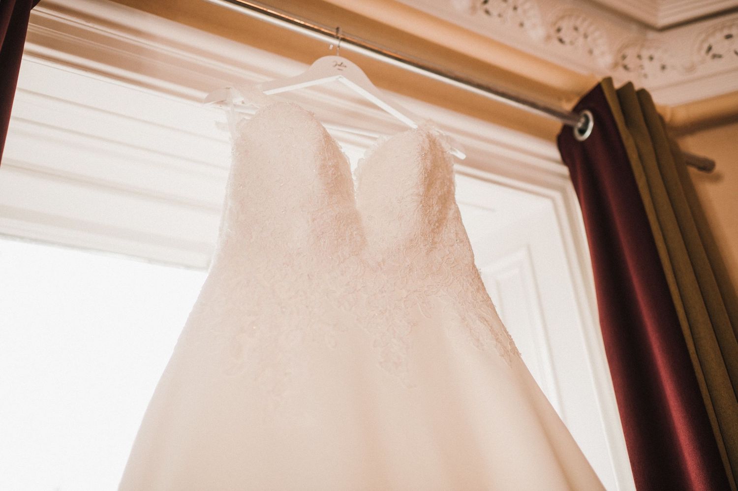 A white wedding dress hangs elegantly in front of a sunlit window with burgundy curtains.