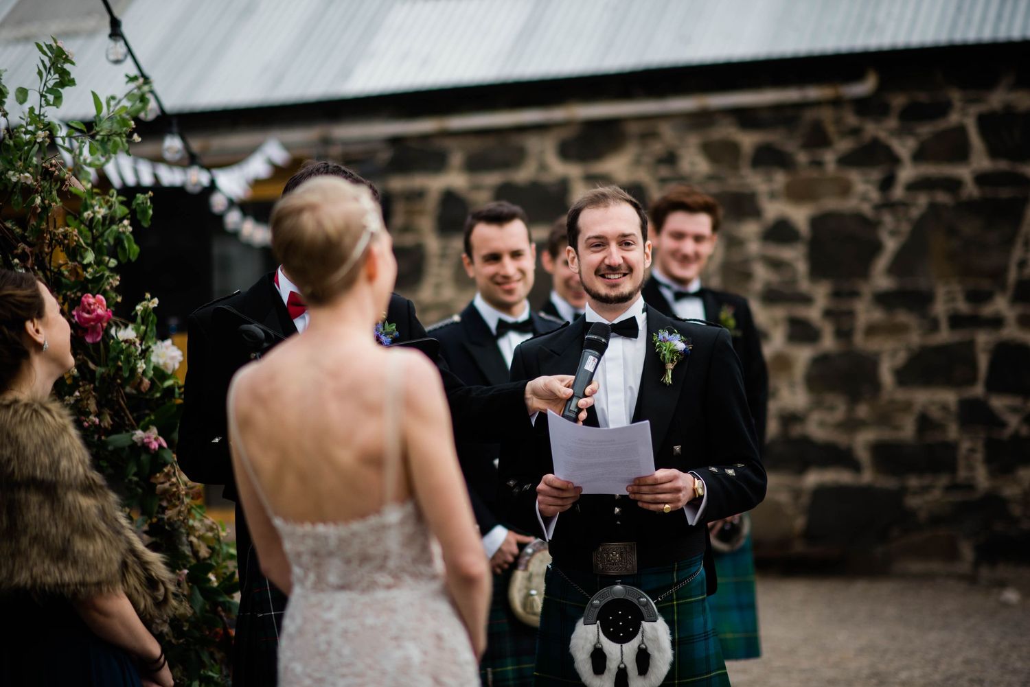 A Scottish wedding ceremony with groomsmen in kilts gathered near a stone wall.