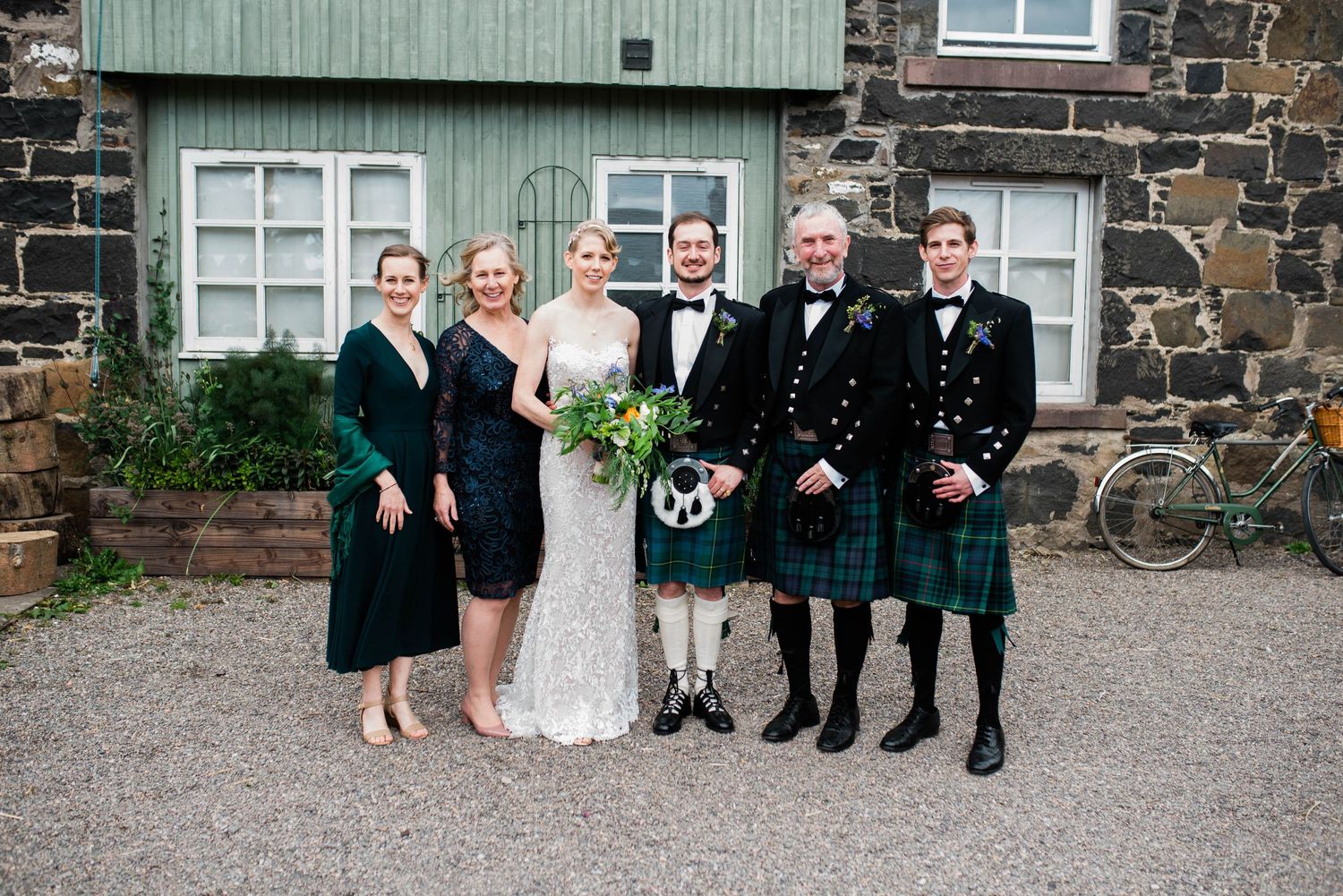 Wedding party poses outside stone building with traditional Scottish kilts and formal attire.