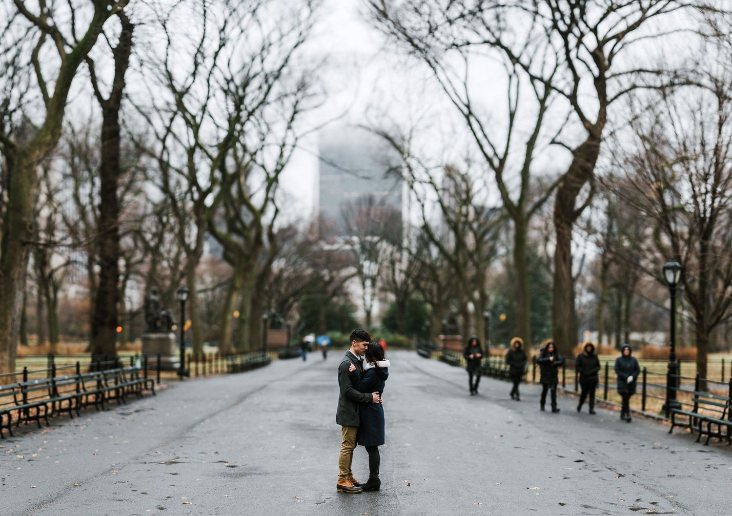 A couple embraces in the distance on a Central Park path with other visitors visible.