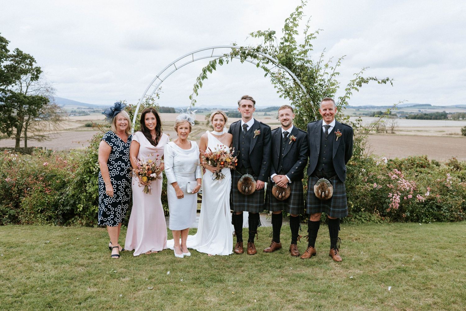 Wedding party poses under floral arch with men wearing traditional Scottish kilts.