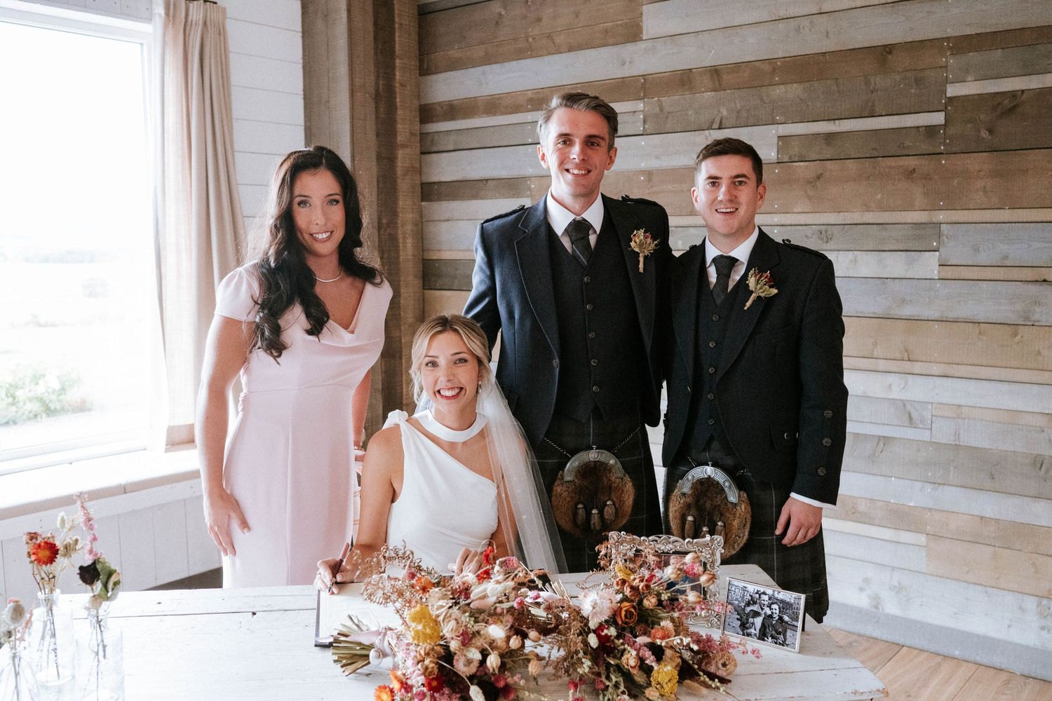 Wedding party gathered in rustic wooden-paneled reception venue with floral decorations.