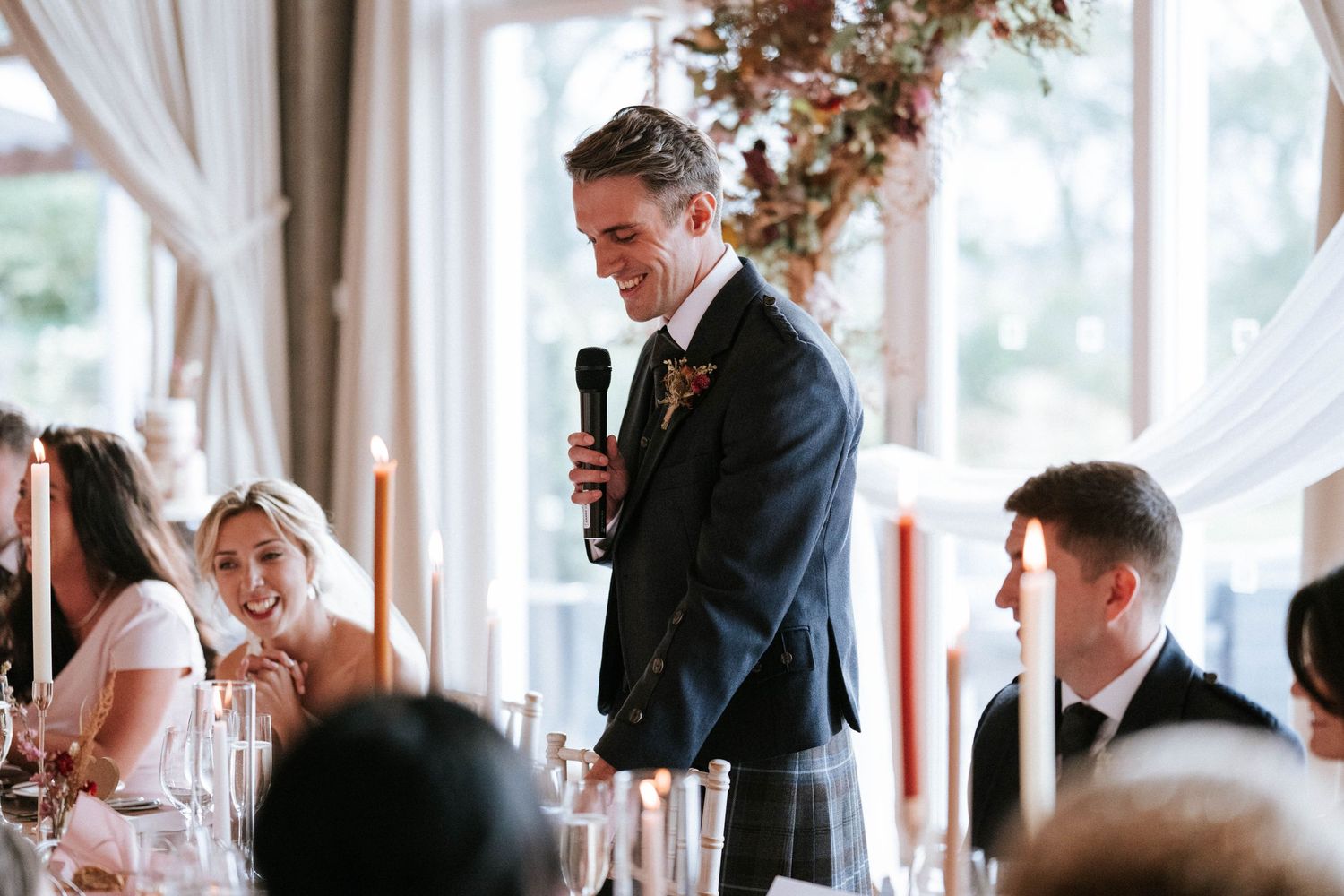 Wedding guests seated at elegant tables in a bright sunlit room with white drapes and autumn decor.