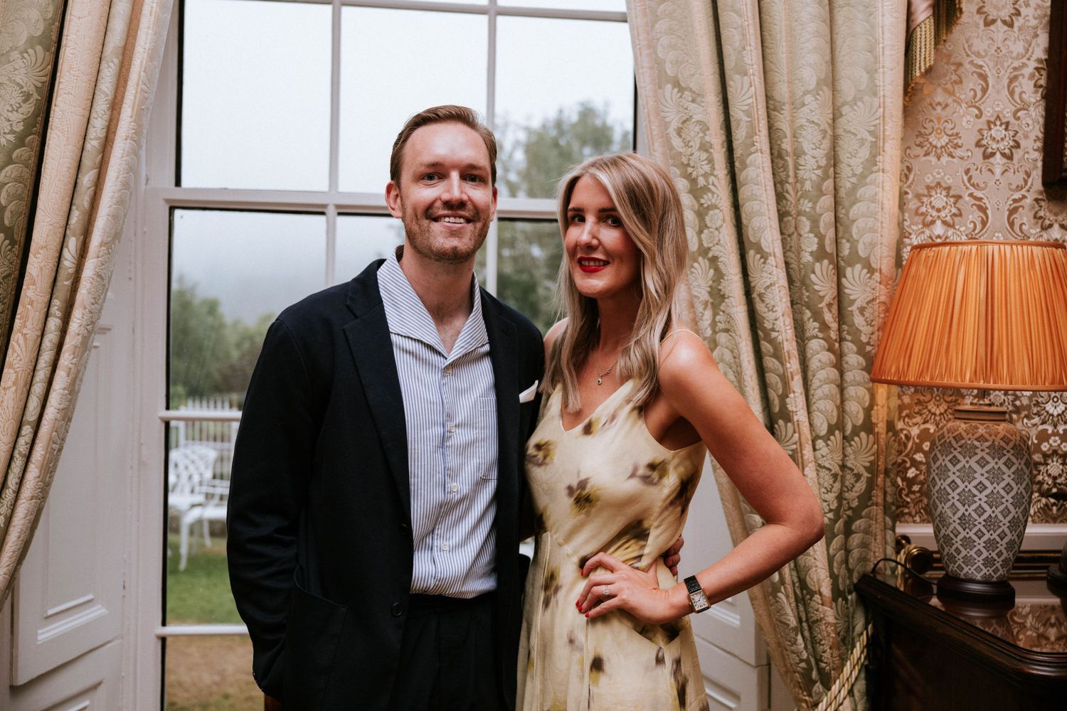 Couple shares a candid moment by large windows in an elegant room.