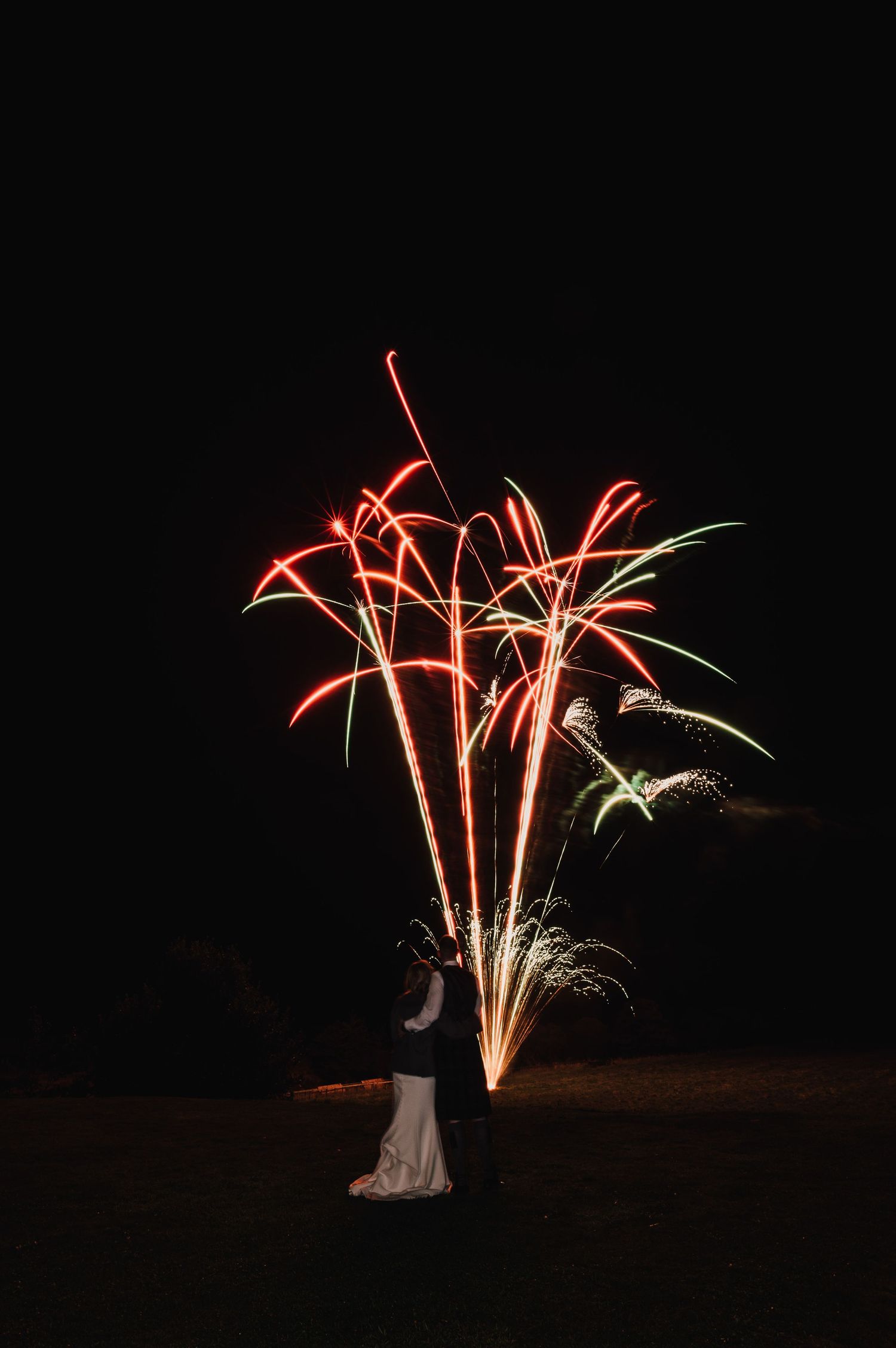 Red and green fireworks burst in night sky above silhouetted figures on grassy field.