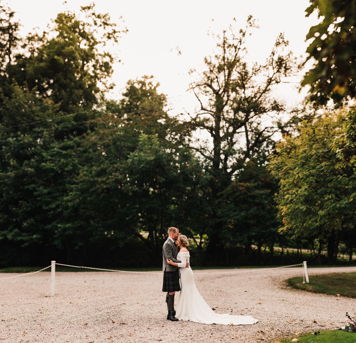 A couple shares an intimate moment on a gravel path surrounded by lush trees at sunset.