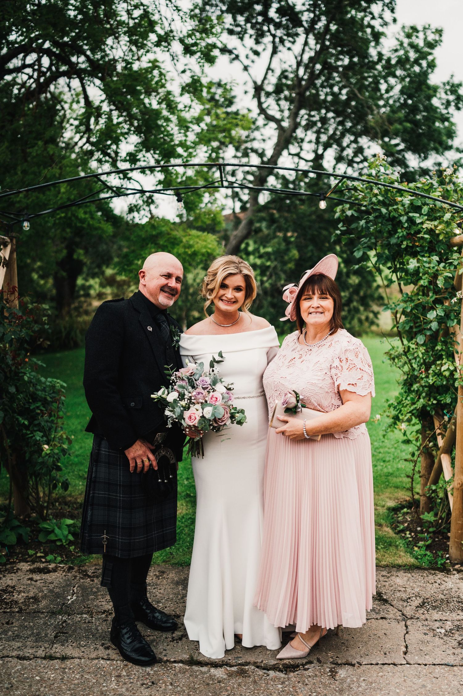 Wedding party members in pink dresses and kilts pose together in a lush garden setting.