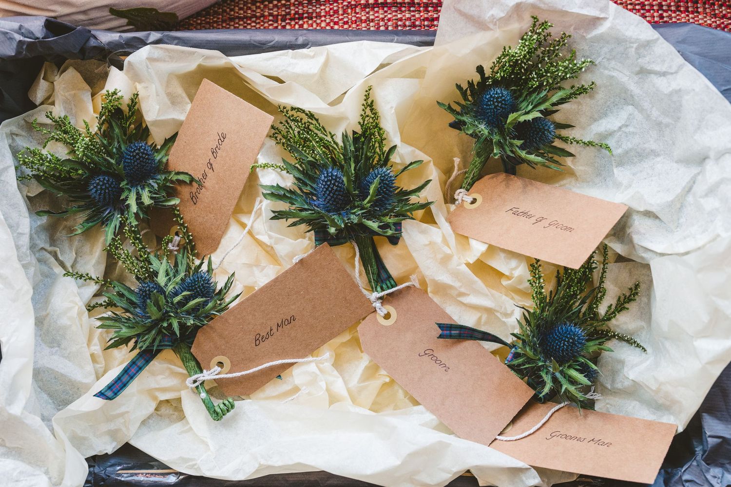 Wedding boutonnieres with blue thistle and greenery laid out on white tissue paper.
