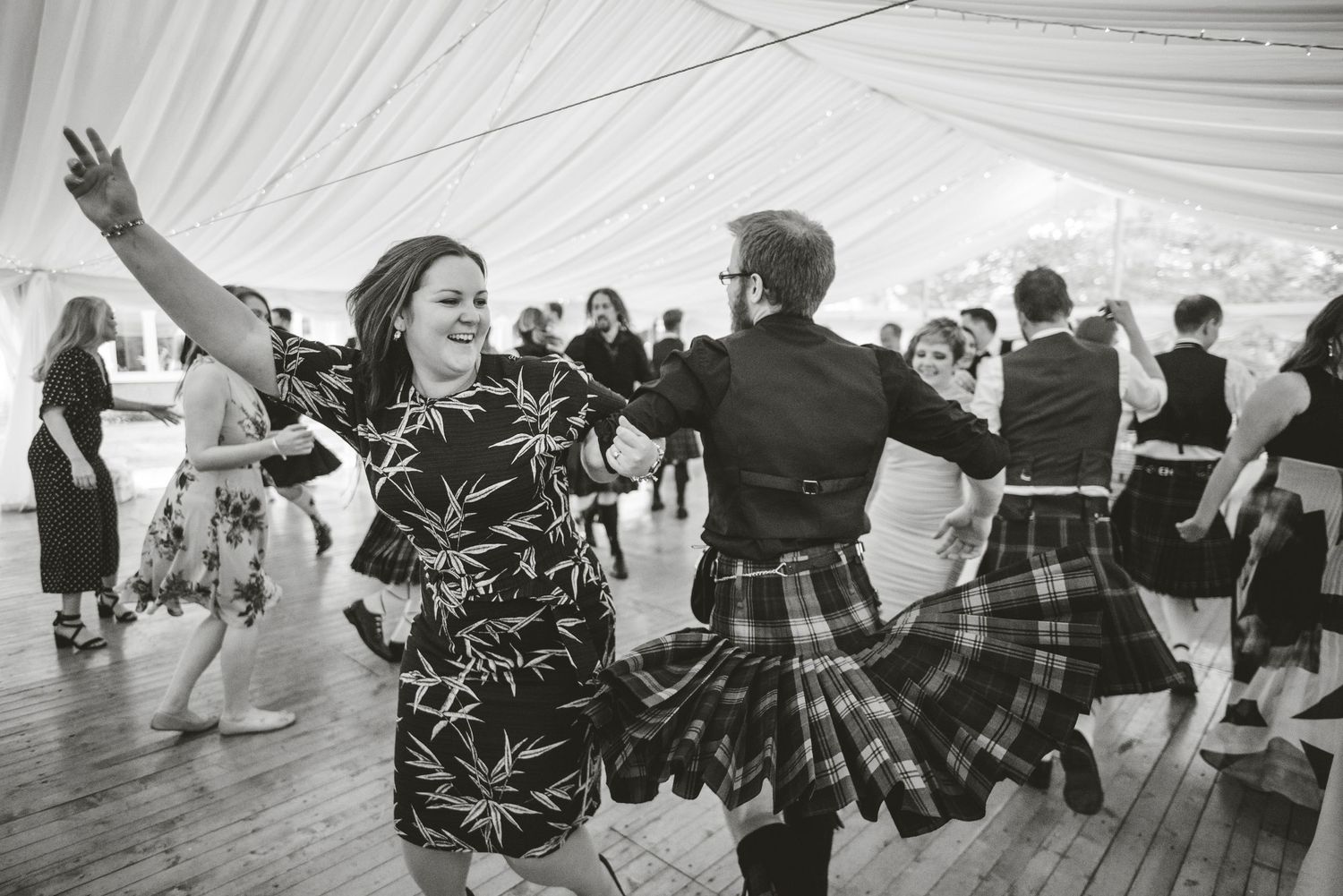 Wedding guests dancing and celebrating under a white tent at an outdoor reception.