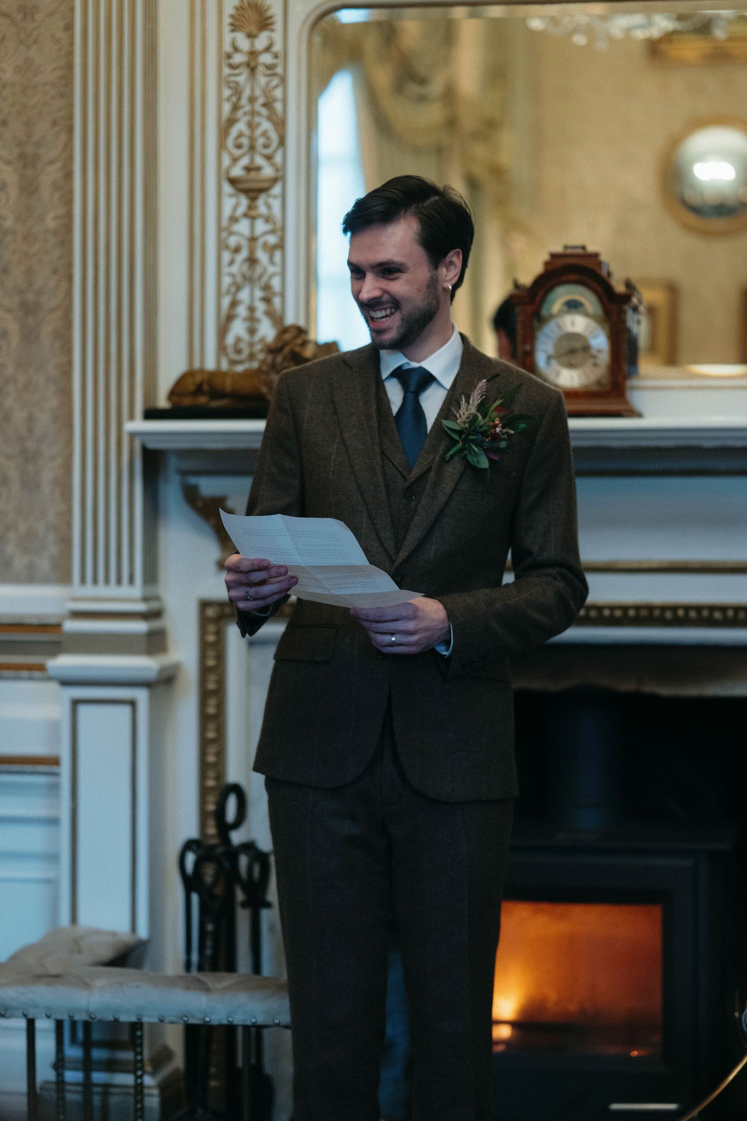 A person in a brown suit reads from papers while standing by an ornate fireplace in a formal room.