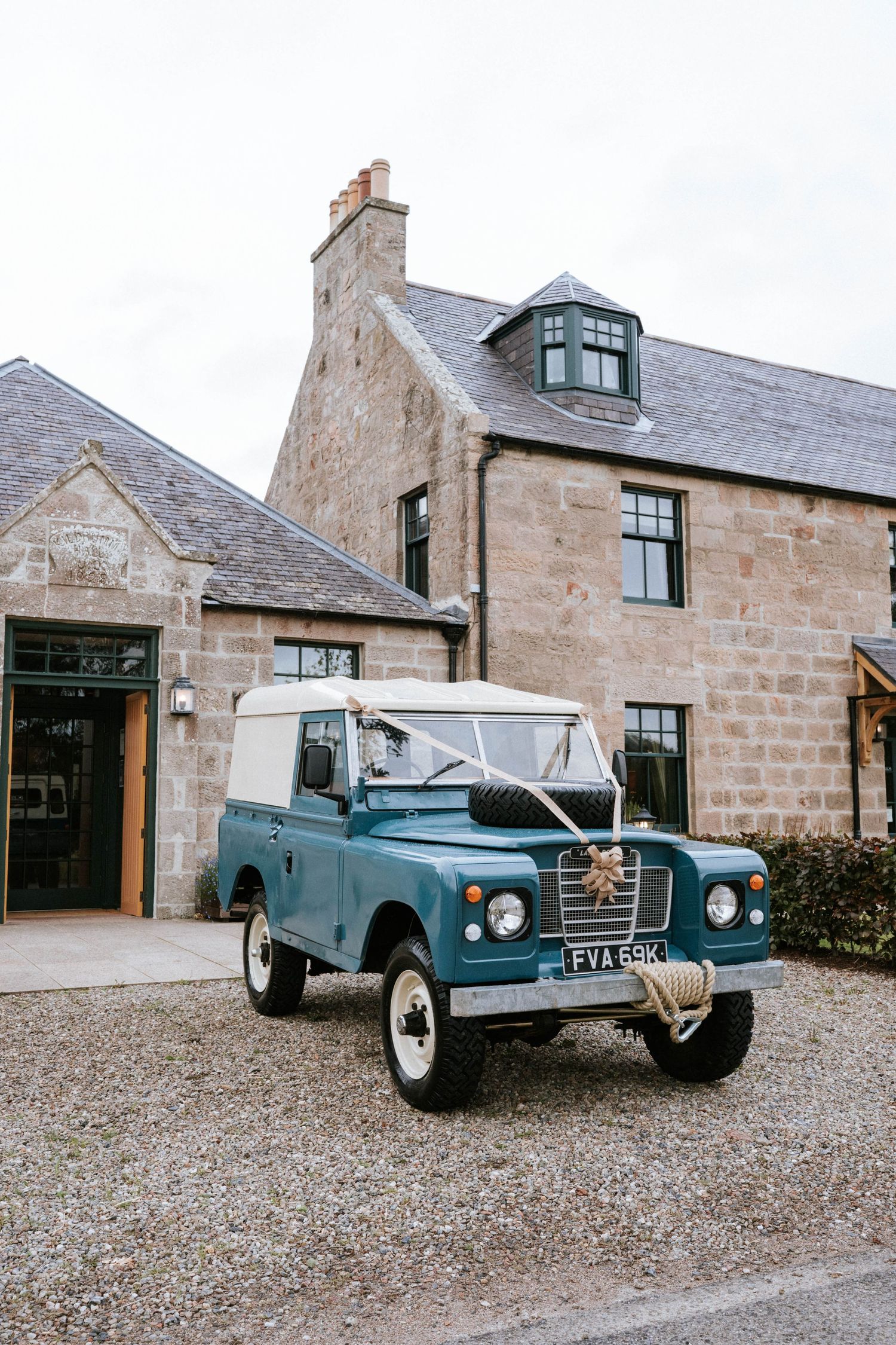 Classic blue vintage Land Rover parked in front of a rustic stone building with gravel driveway.