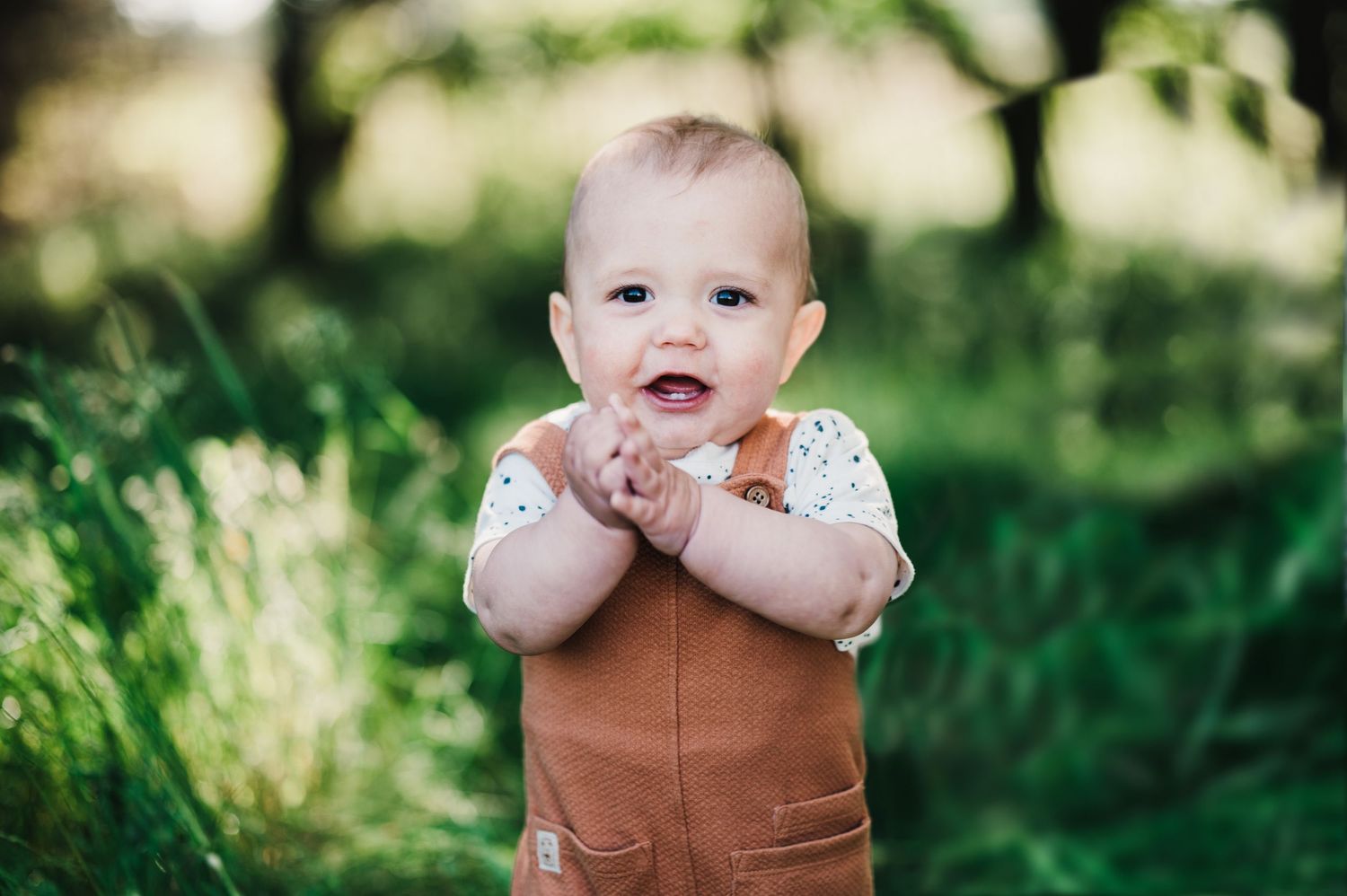 A small child in a brown vest smiles while playing outdoors in a lush green garden setting.