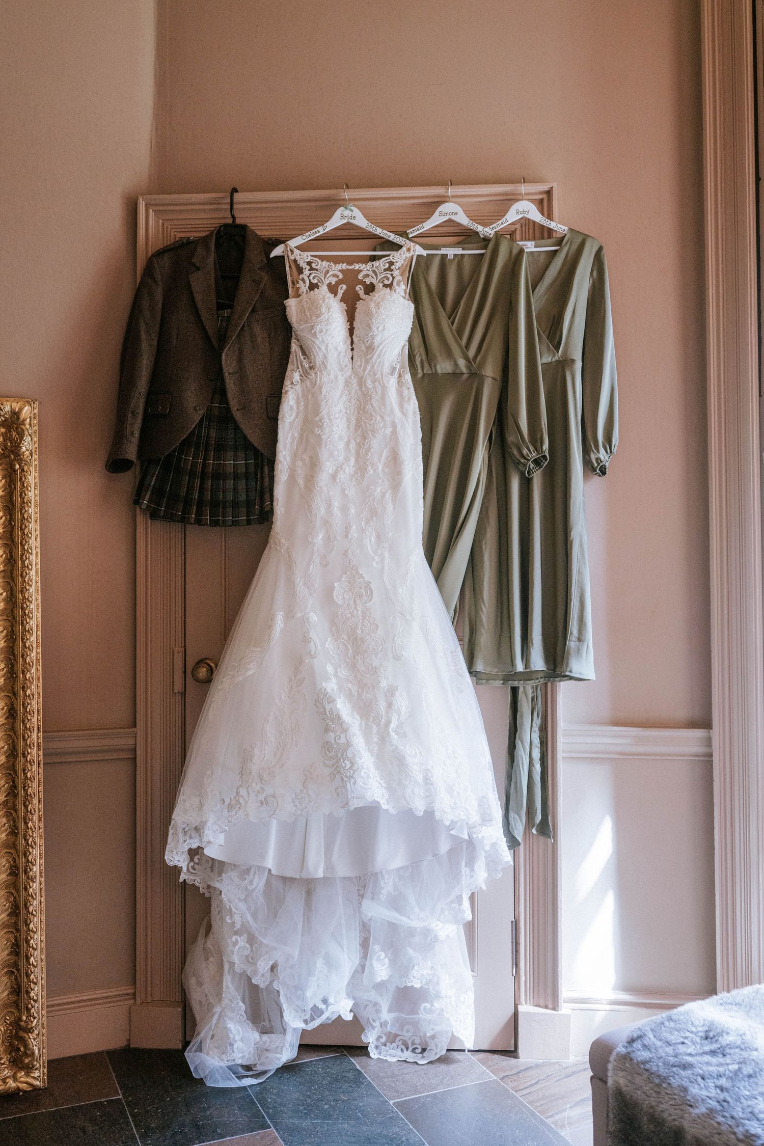 Wedding dress hanging between green bridesmaid dresses and dark jacket on door.