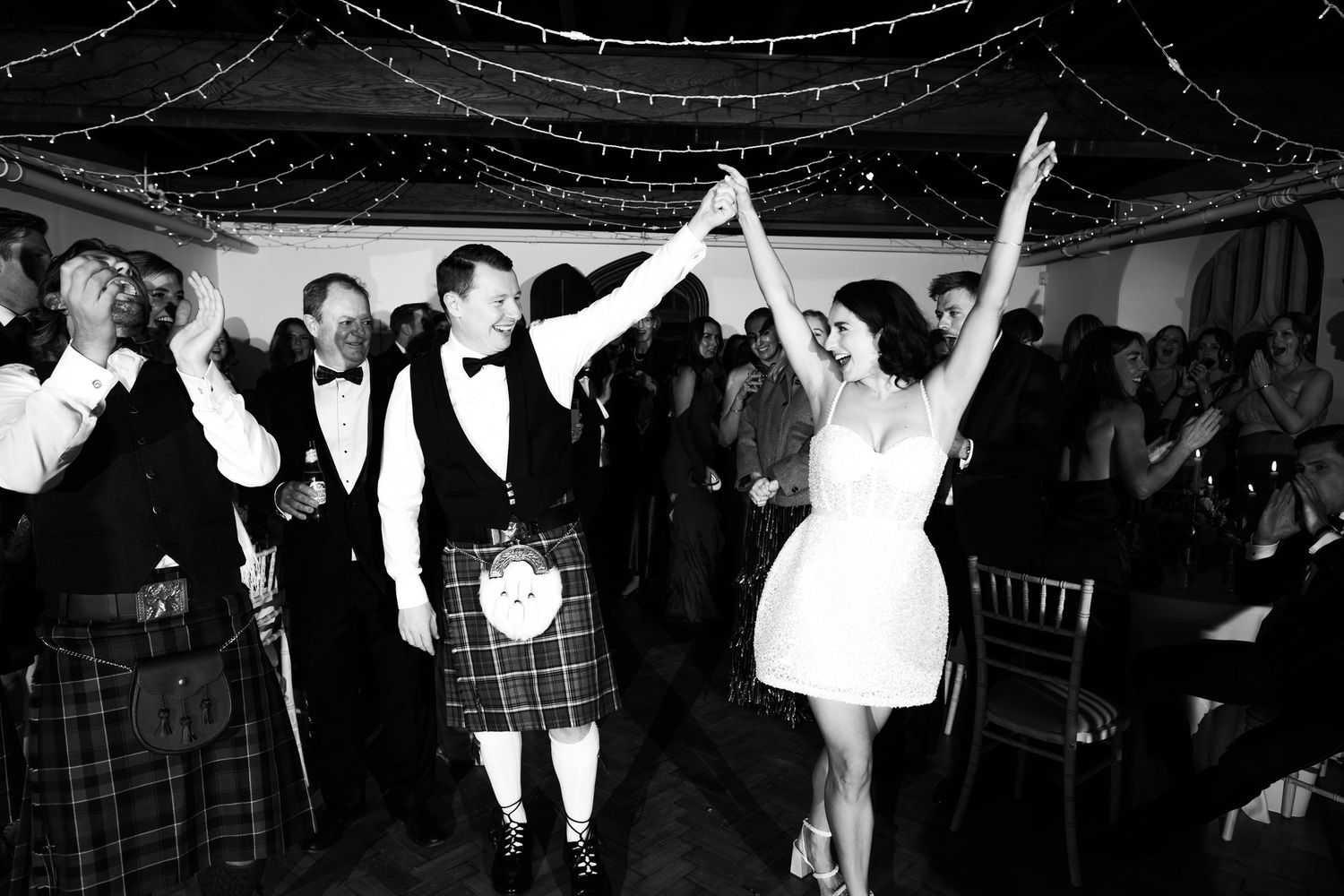 Wedding guests dance enthusiastically on a dimly lit dance floor beneath decorative string lights.