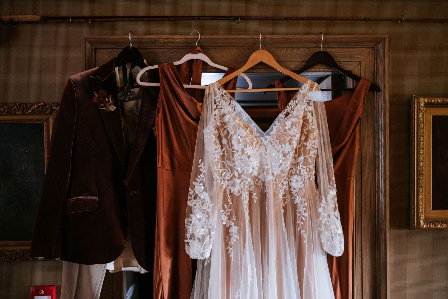 White lace wedding dresses hanging on wooden hangers against dark wall with warm lighting.