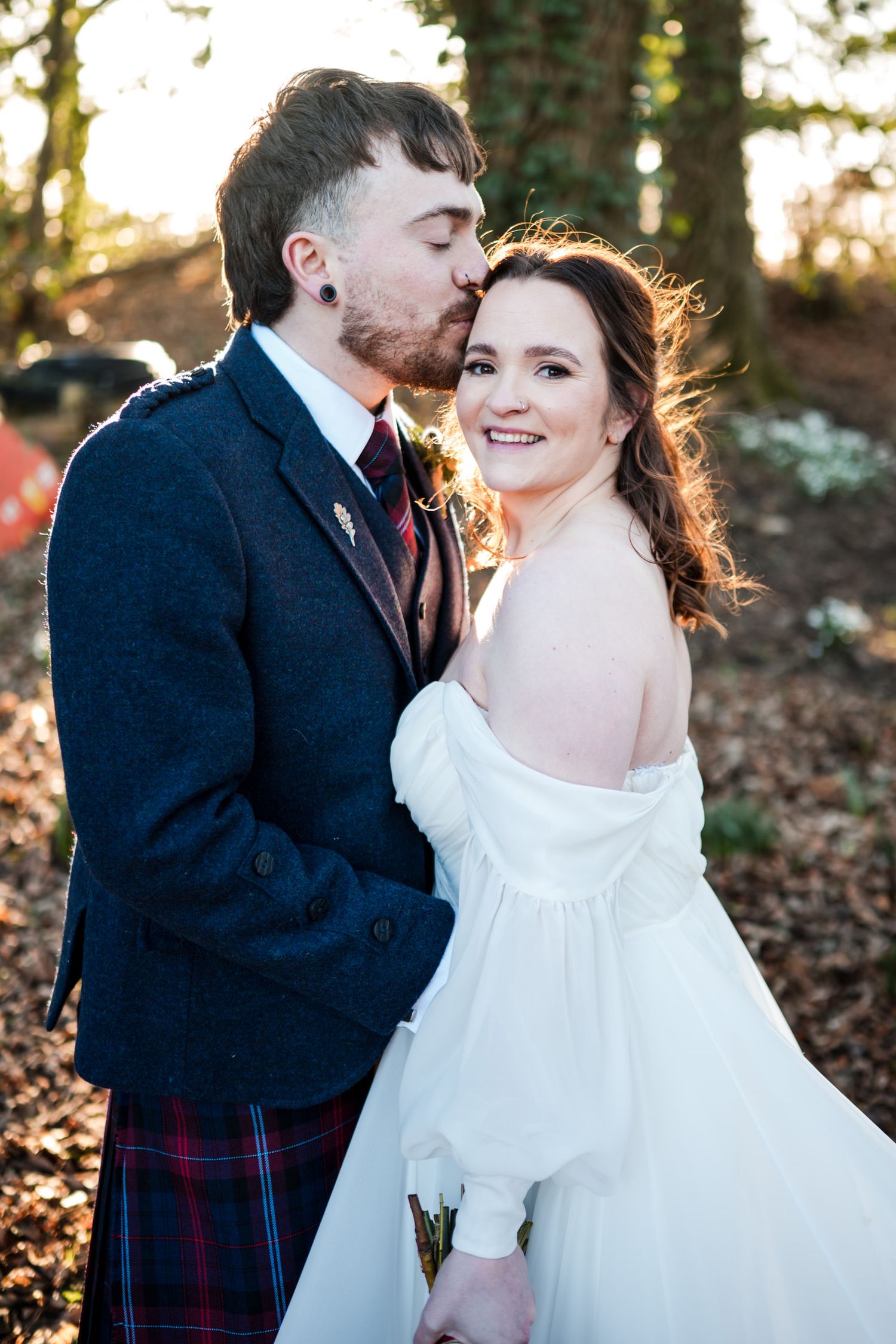 A romantic winter wedding couple embraces in a forest setting with fallen leaves and golden sunlight.