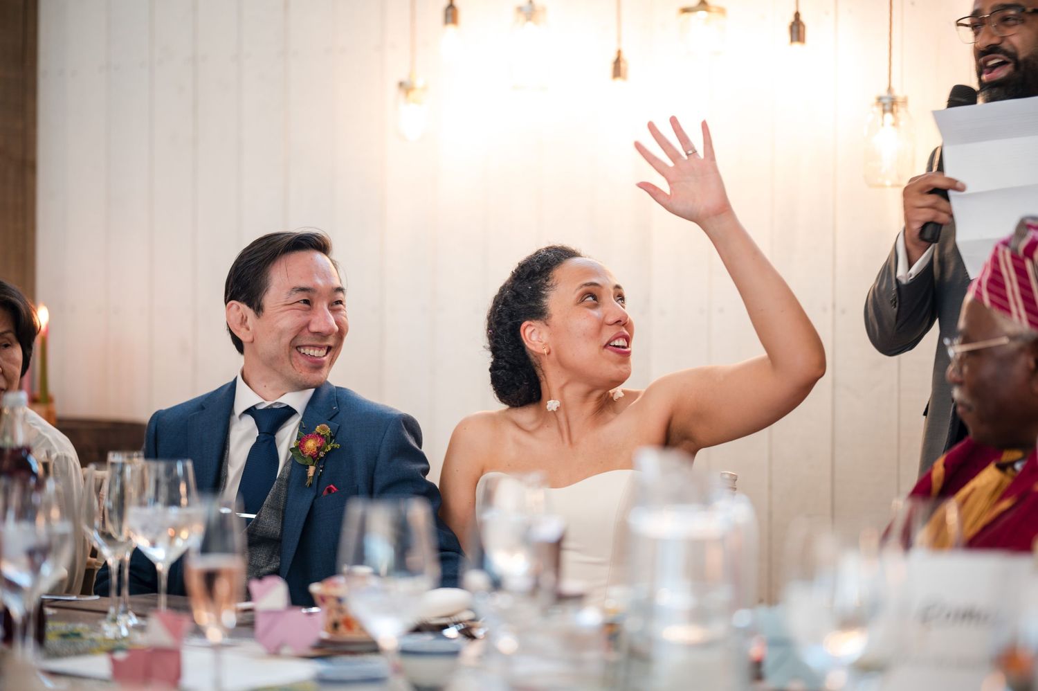 Wedding reception guests laughing and celebrating at indoor venue with pendant lighting.