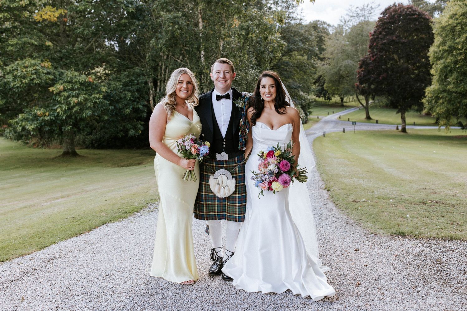 Three people in formal attire including Scottish kilt pose on garden path.